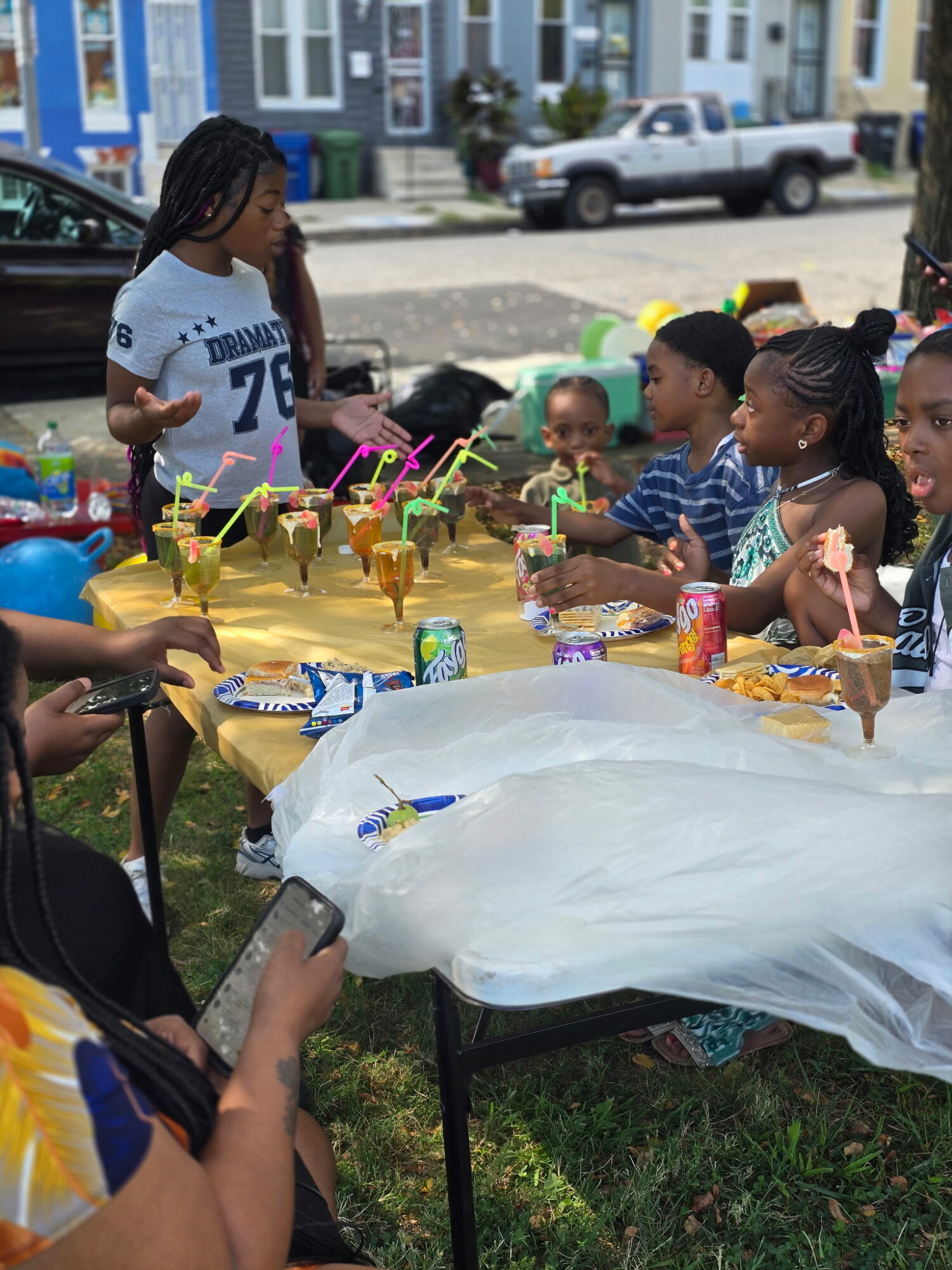 Children and adults gather around a table outdoors with colorful decorations and drinks, celebrating a festive event on a sunny day.
