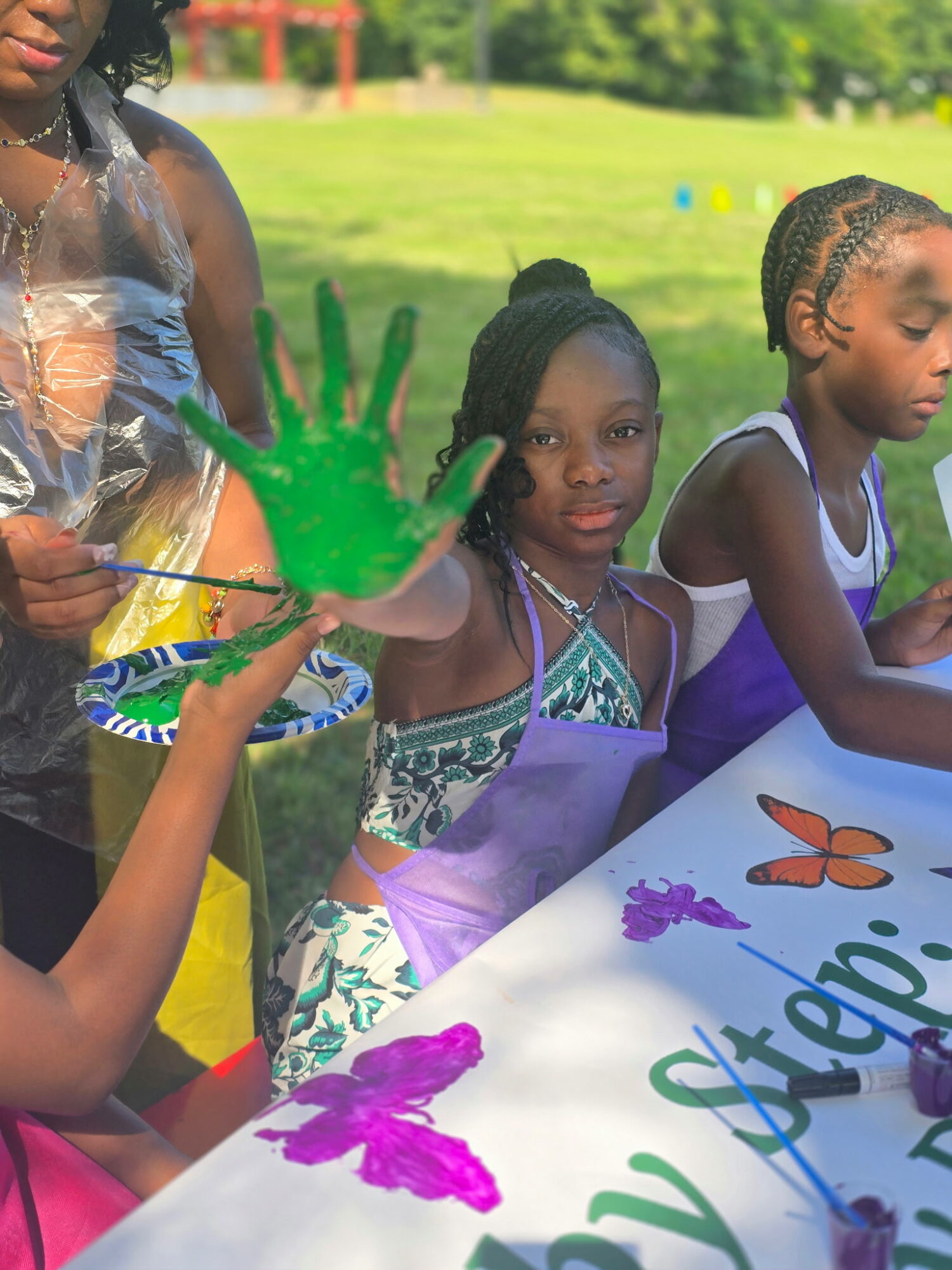 Three children sitting at a table outdoors, one with a painted green handprint on their palm, colorful butterfly and flower decorations, and a grassy background.