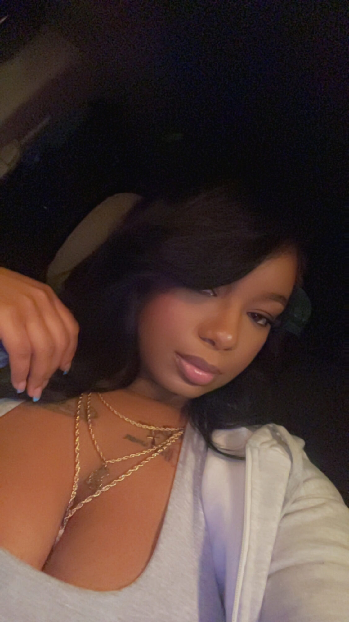 Woman with dark hair and makeup, wearing layered necklaces and a light-colored top, sitting in a car at night.
