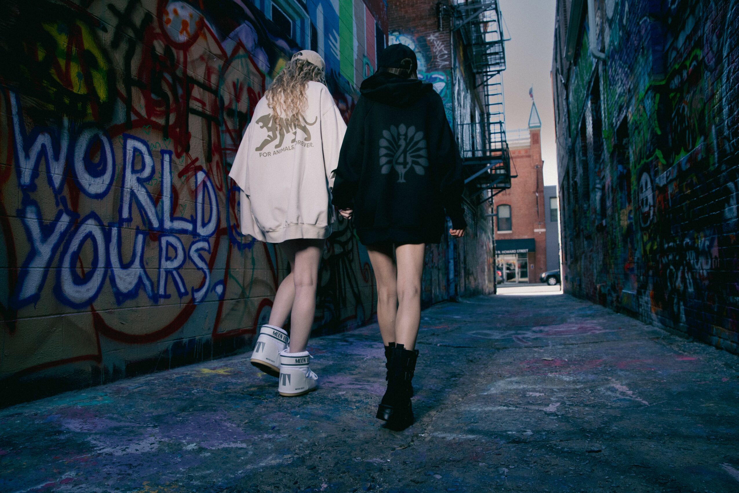 Two people walk through a graffiti-covered alleyway with colorful murals on walls, wearing casual clothing and boots.