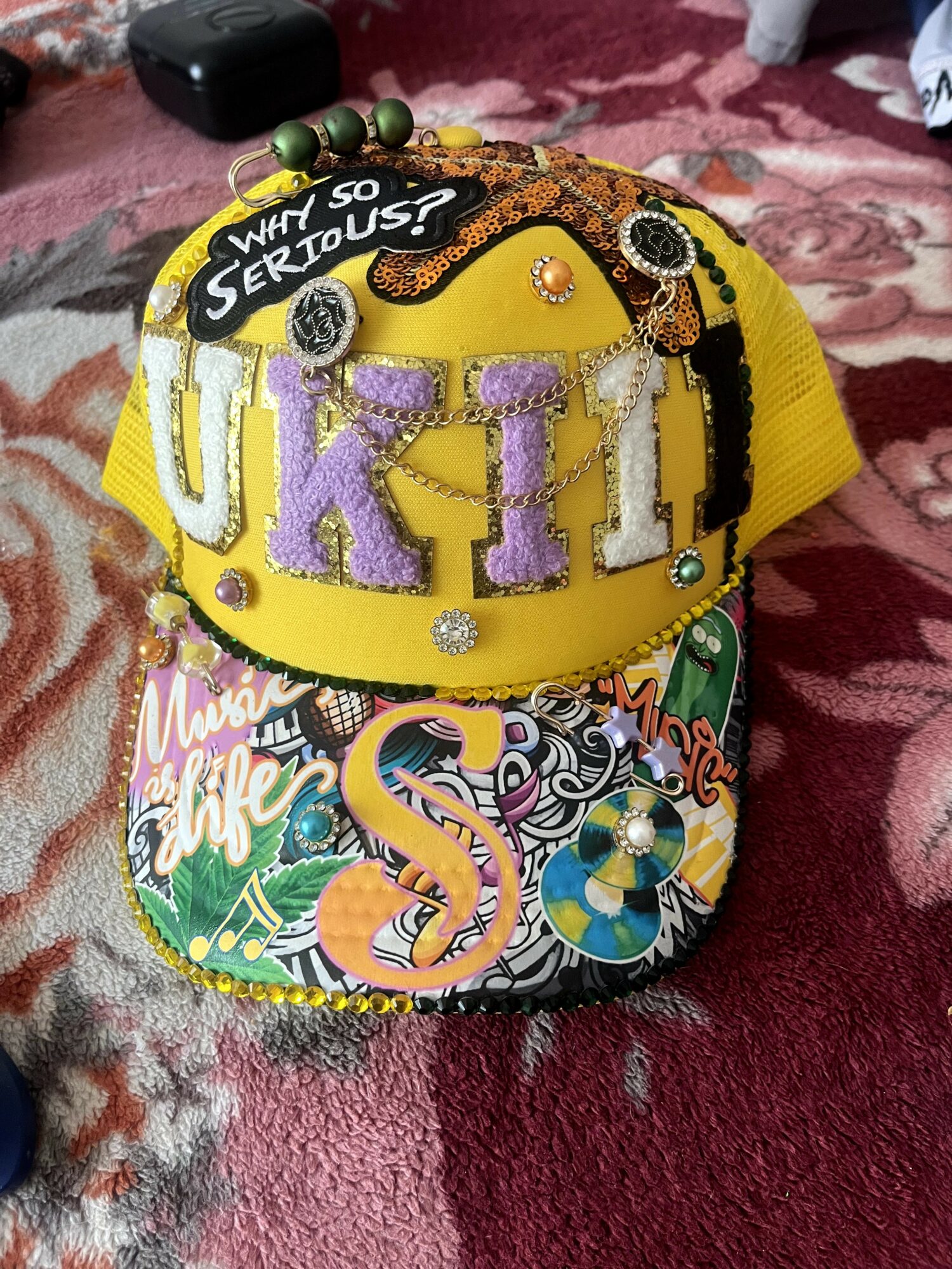Colorful decorated cap with purple letters, beads, and a patterned fabric base, placed on a pink and red textured surface.