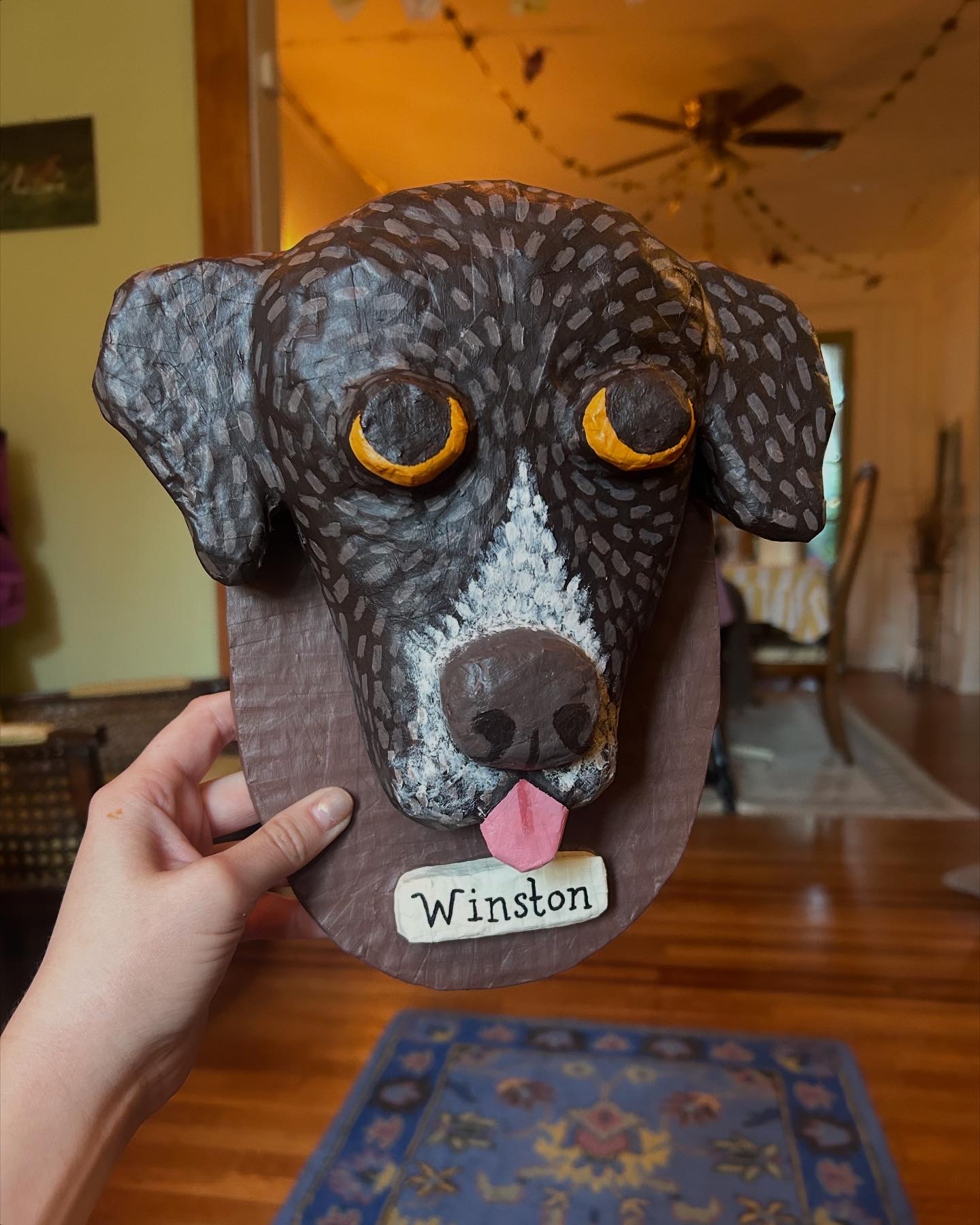 Hand holding a paper mask of a dog's face with yellow eyes and a pink tongue, labeled 'Winston'.