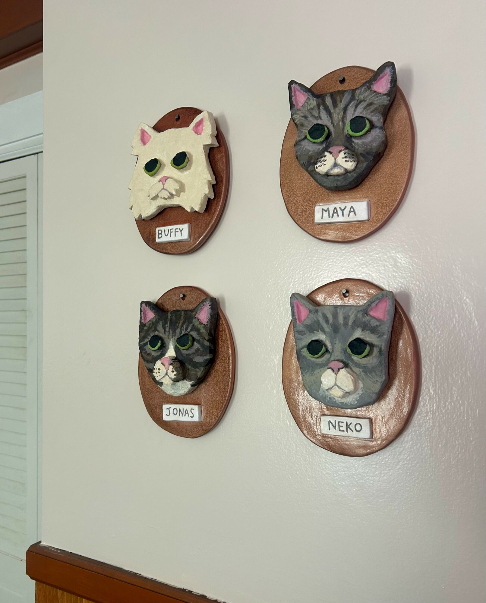 Four decorative wall plaques with cat faces and names: Buffy, Maya, Jonas, Neko, on a plain wall.