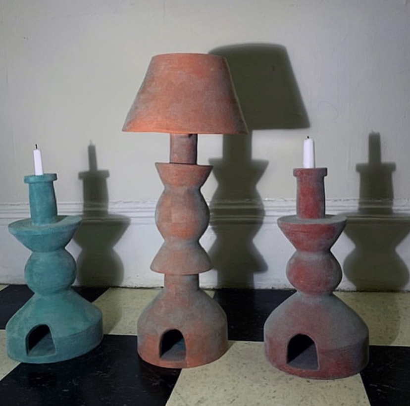 Three candle holders with candles, designed as abstract structures, cast shadows on a wall. One has a lamp shade. 20 words.