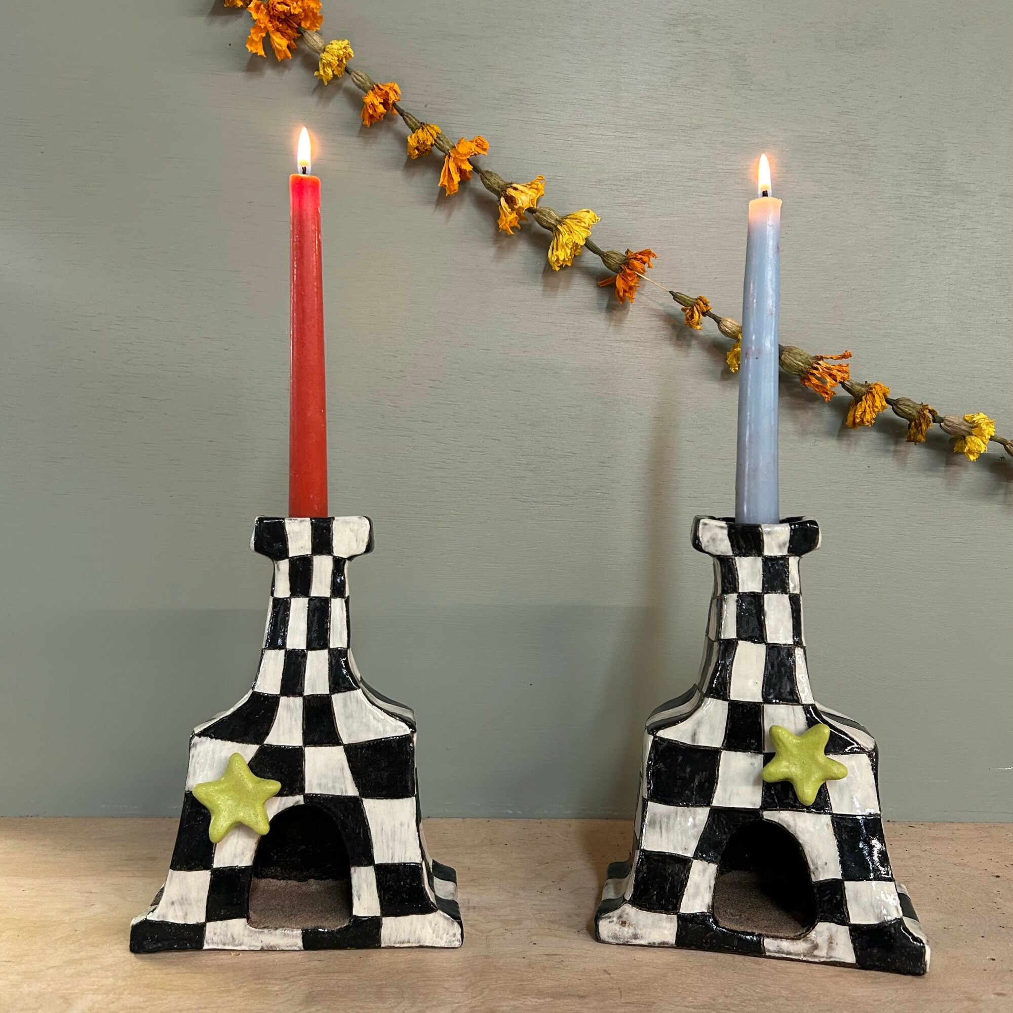 Two checkered candle holders with lit candles, one red and one blue, on a wooden surface with a yellow flower garland above. Word count: 24