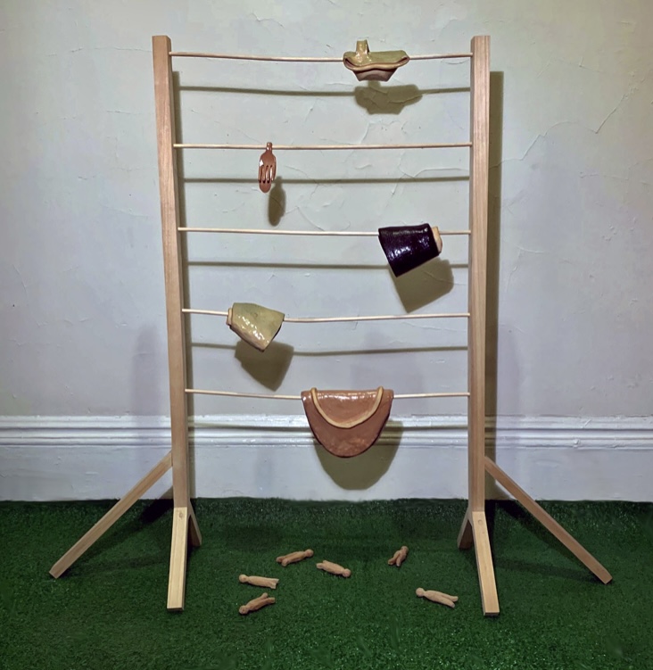 Wooden ladder with toy objects hanging on horizontal bars, on green carpet, against a light-colored wall. Several chalk pieces are on the floor.