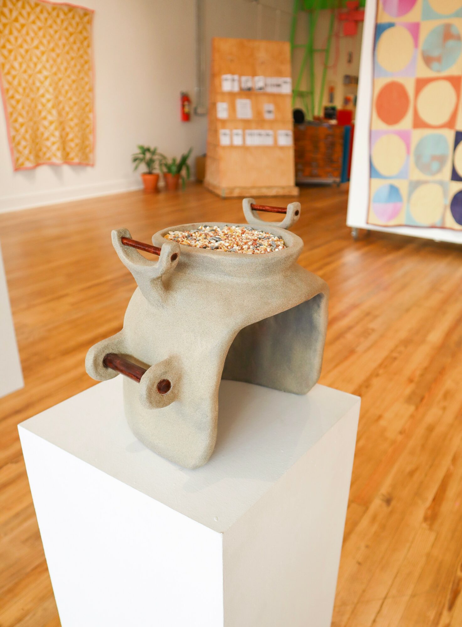 Ceramic sculpture of a stylized animal with a hollow top, handles, and a tail, displayed on a white pedestal in an indoor space.