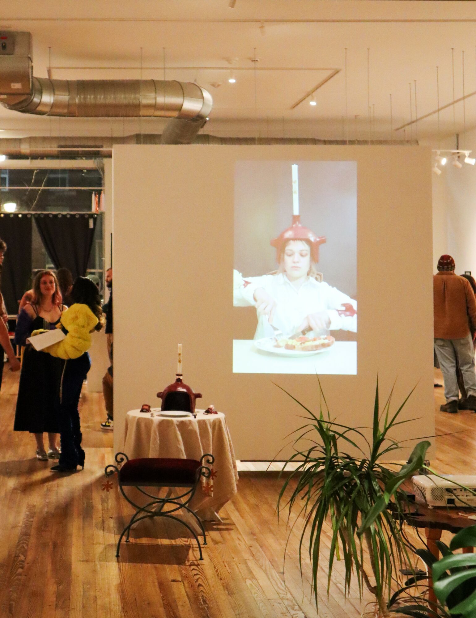 Projection of a person wearing a red hat on a white wall in an indoor space with people and plants.