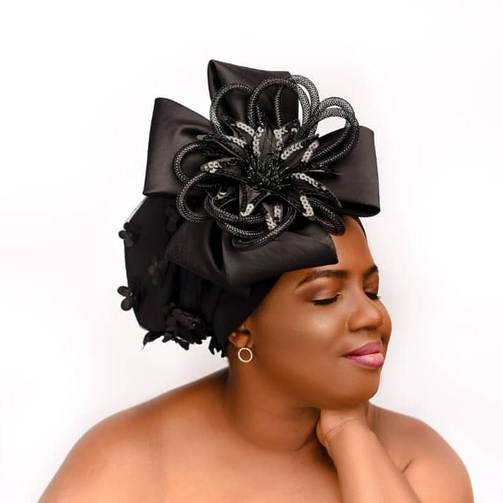 Woman with elaborate black headwrap and earrings, eyes closed, smiling gently, against a white background.