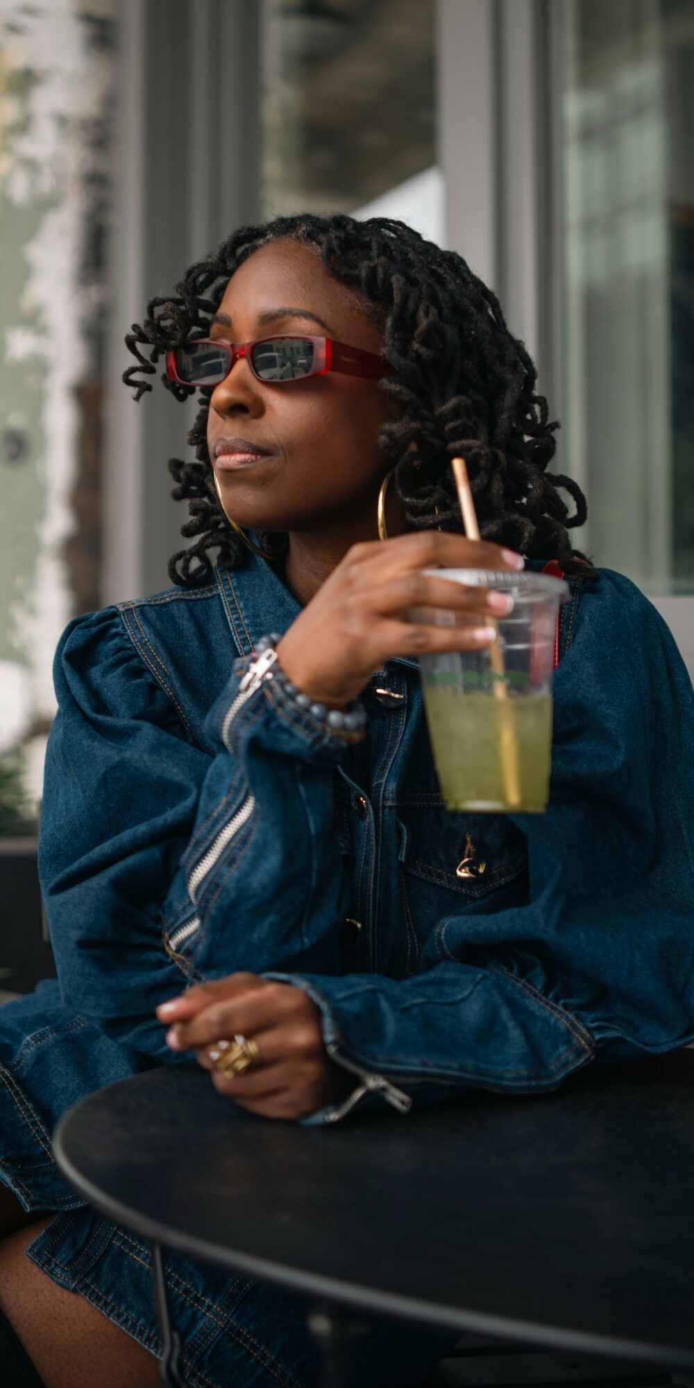 Woman with curly hair and sunglasses holding a drink with a straw, sitting outdoors near a window.