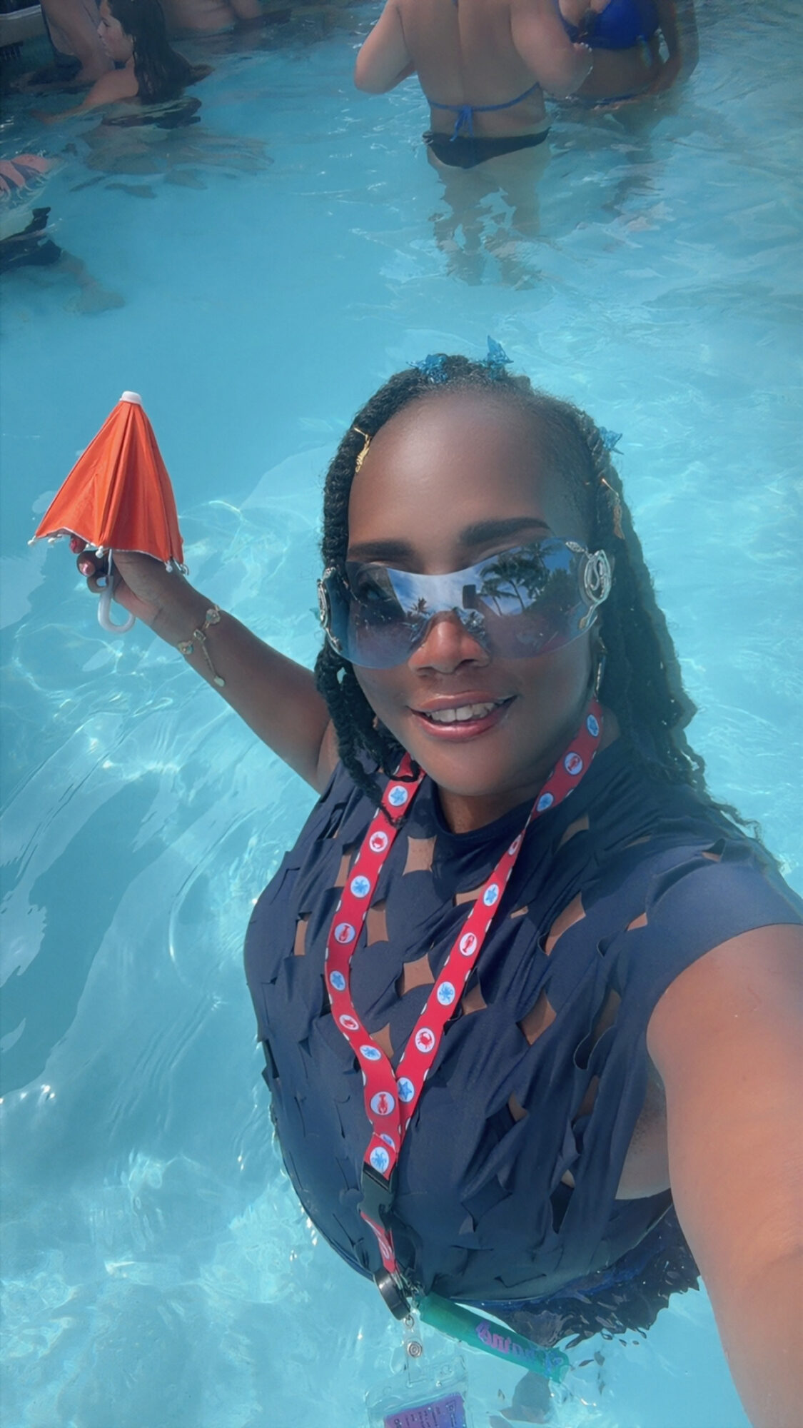 Woman in swimming pool holding orange umbrella, wearing sunglasses and a dark shirt, with other people in the background.