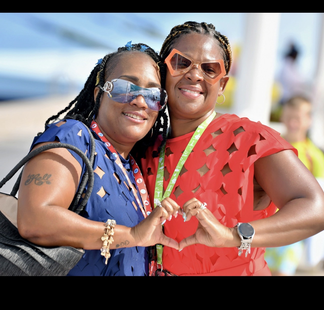Two women smiling, making a heart shape with their hands, outdoors on a sunny day, wearing colorful clothing and sunglasses.
