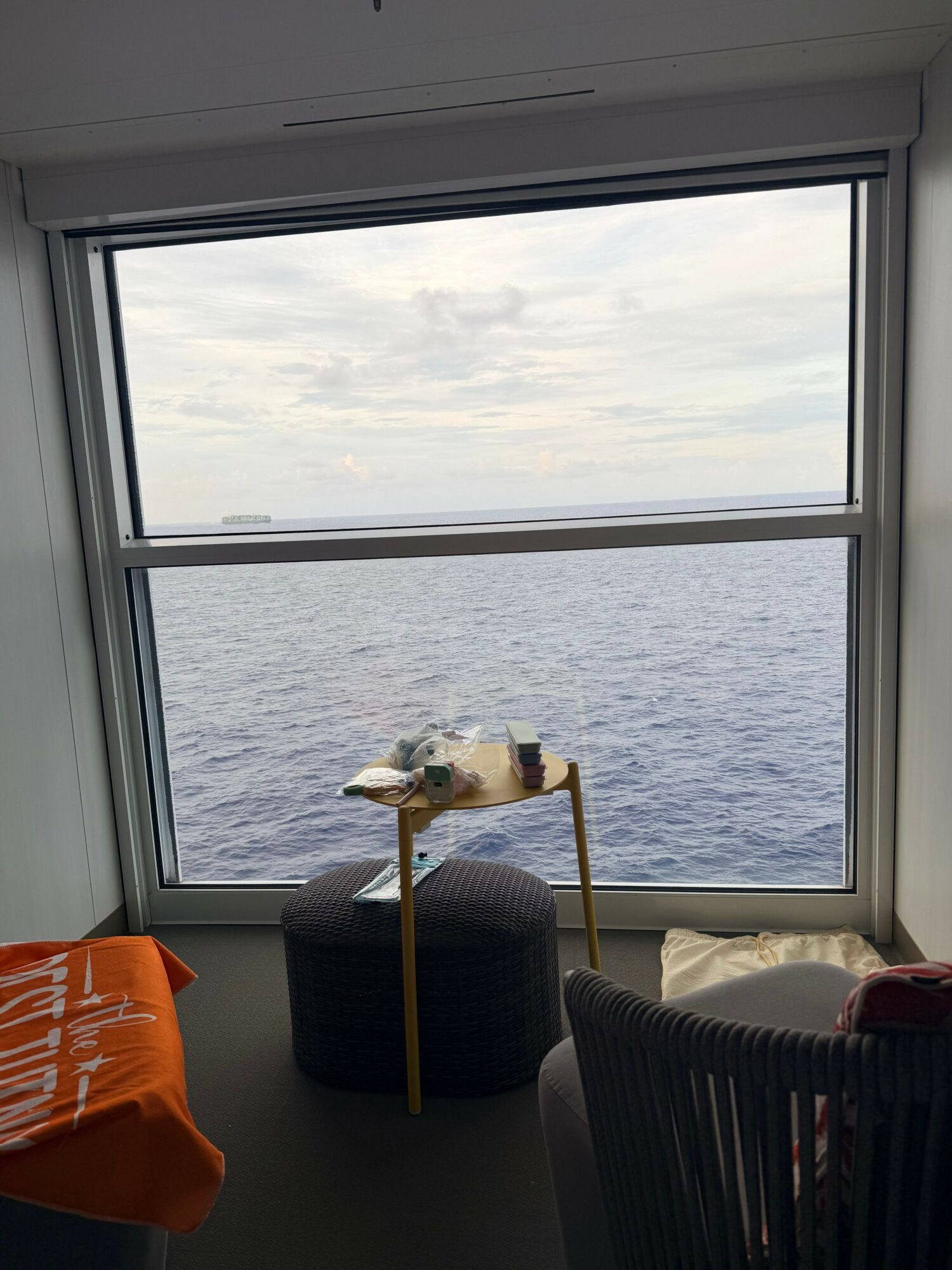 View of a large window overlooking a body of water with a cloudy sky, with a table and chair inside.