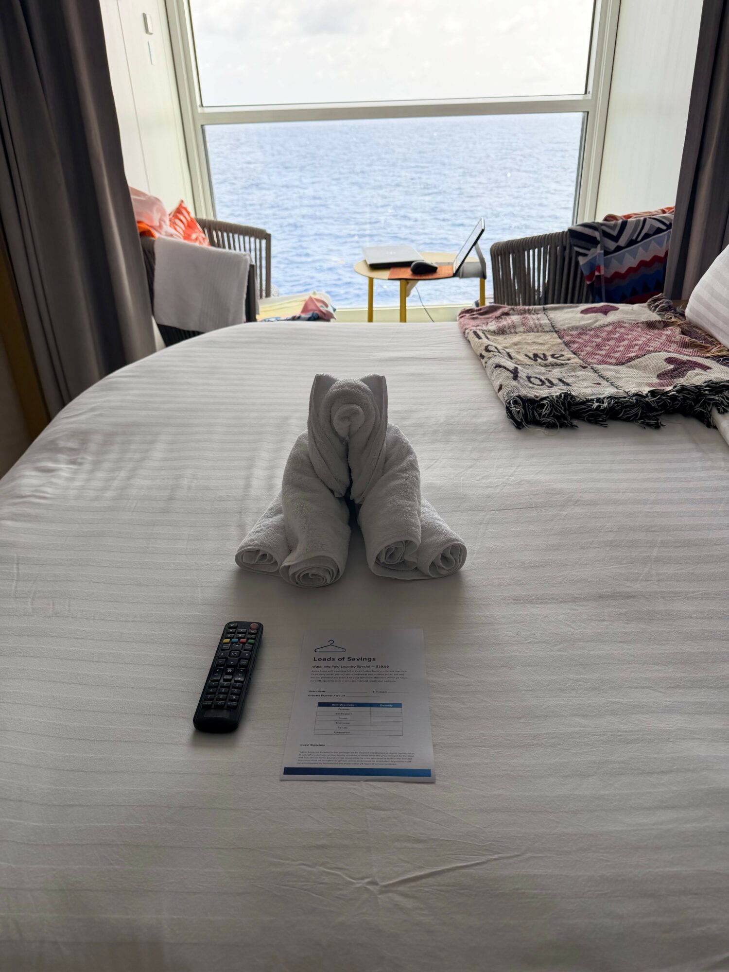 A hotel bed with two rolled towels, a remote control, and a paper on the white bedspread, with a window showing water outside.