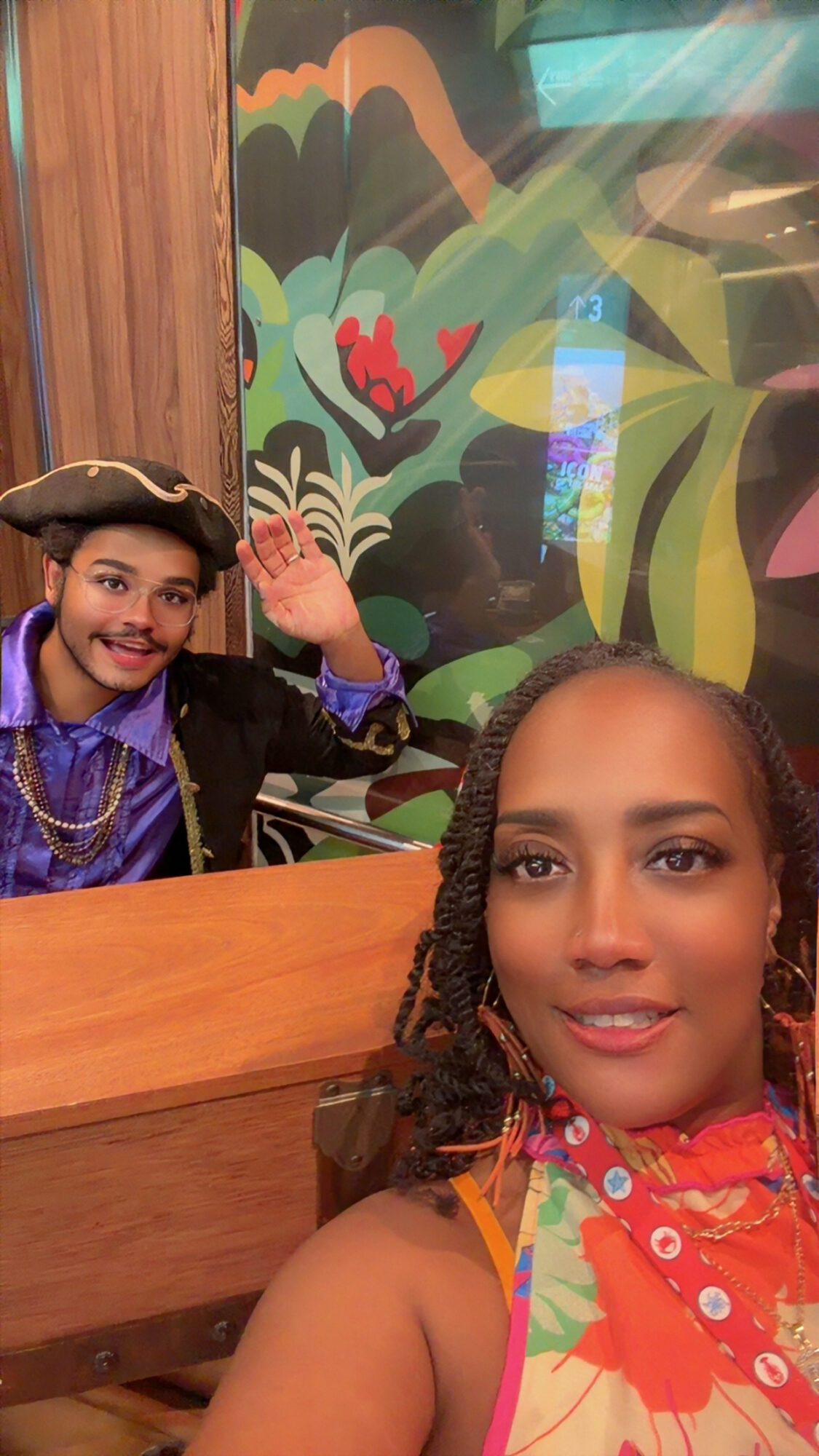 Two people, one in pirate costume and one in colorful clothing, smiling indoors with a colorful mural in the background.