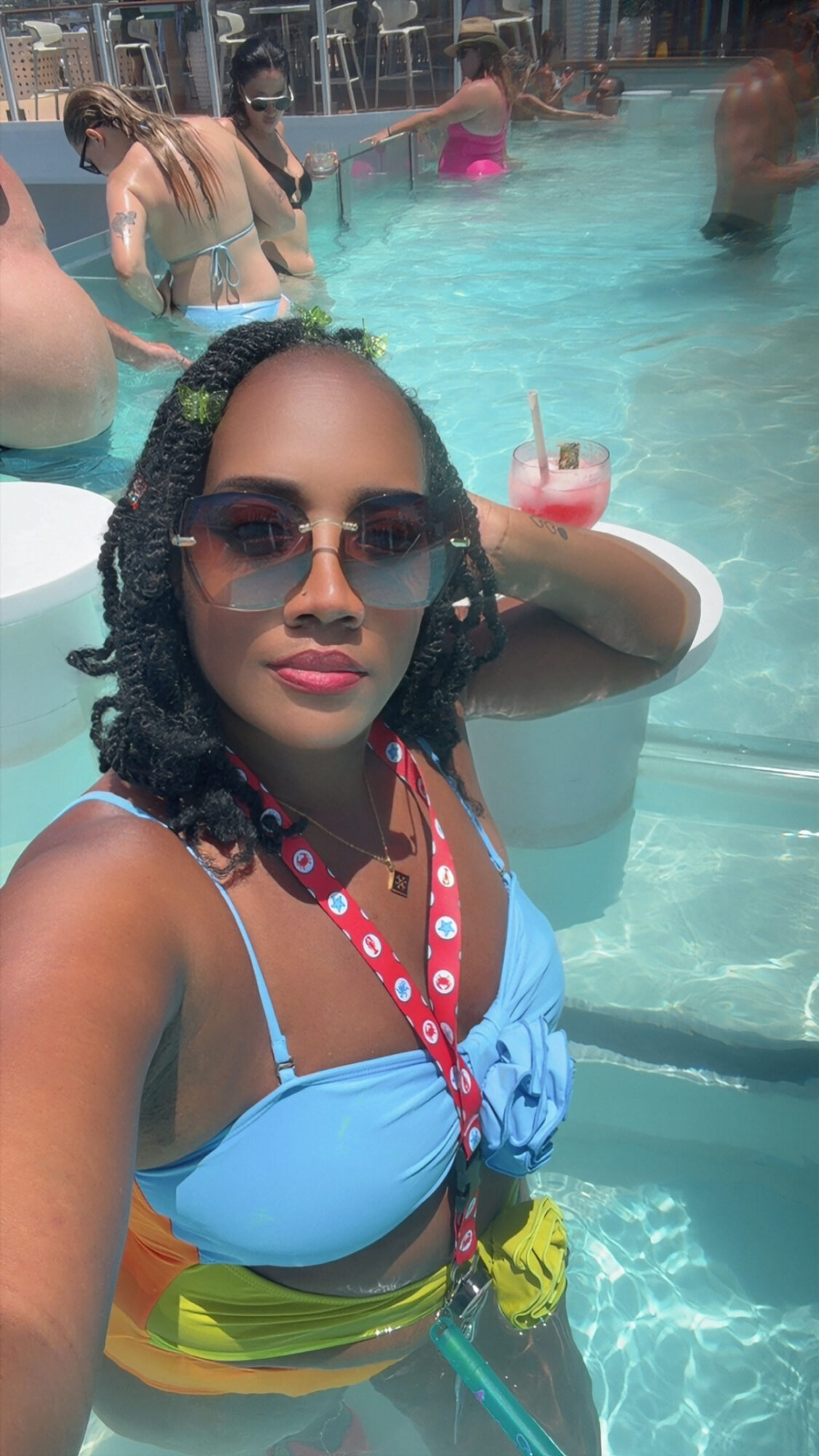 Woman with sunglasses and braided hair in a pool, holding a drink, wearing a blue top and colorful shorts.