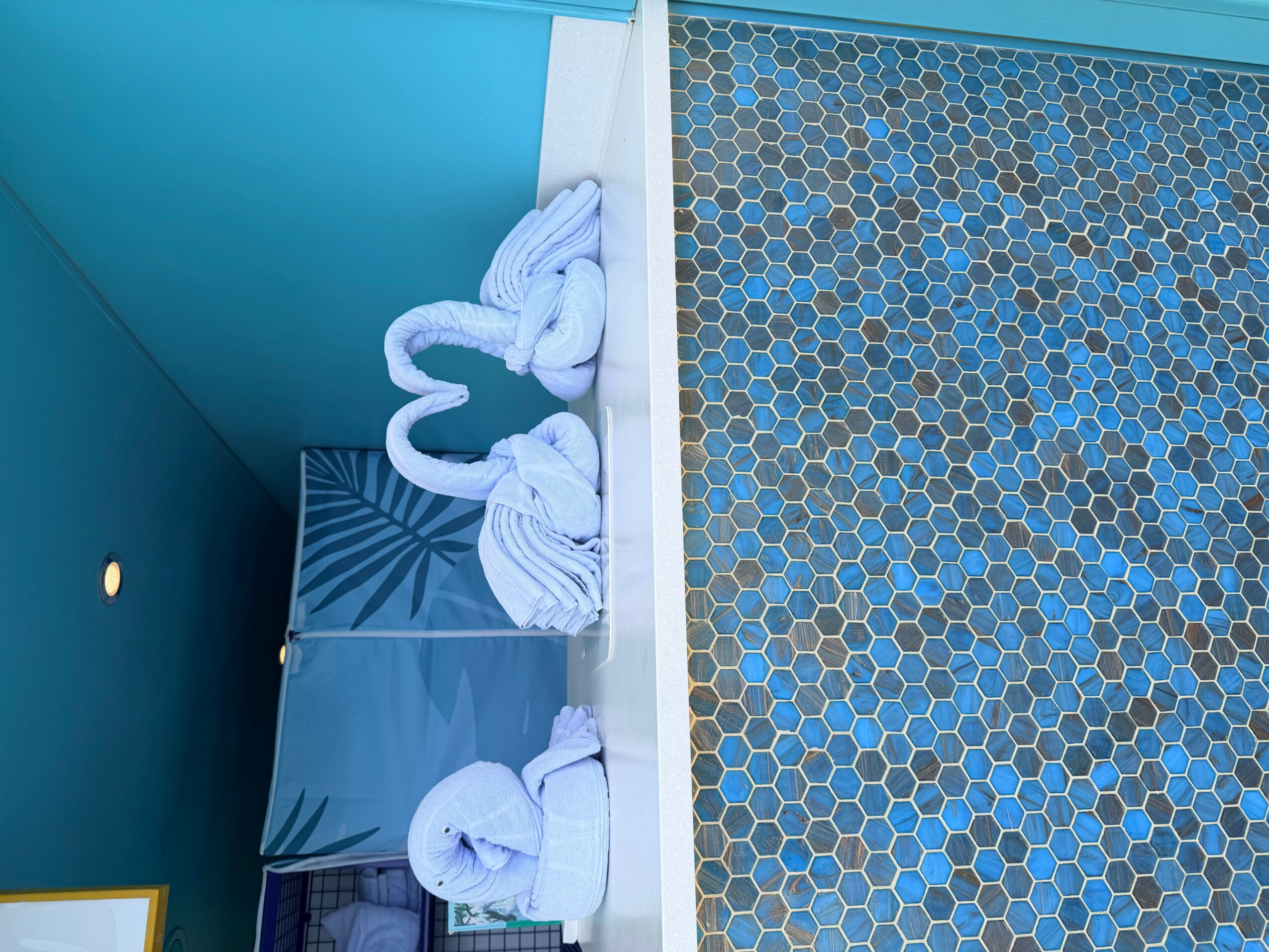 Towels folded into swans on a shelf next to a mosaic-tiled wall, with a blue wall and a small plant in the background.