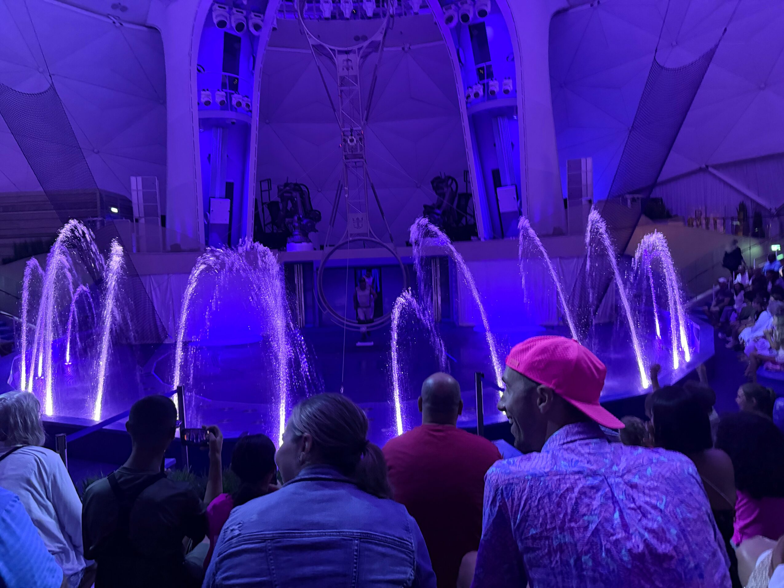 Audience watching a fountain show with purple lighting at an indoor venue, with a futuristic ceiling design.