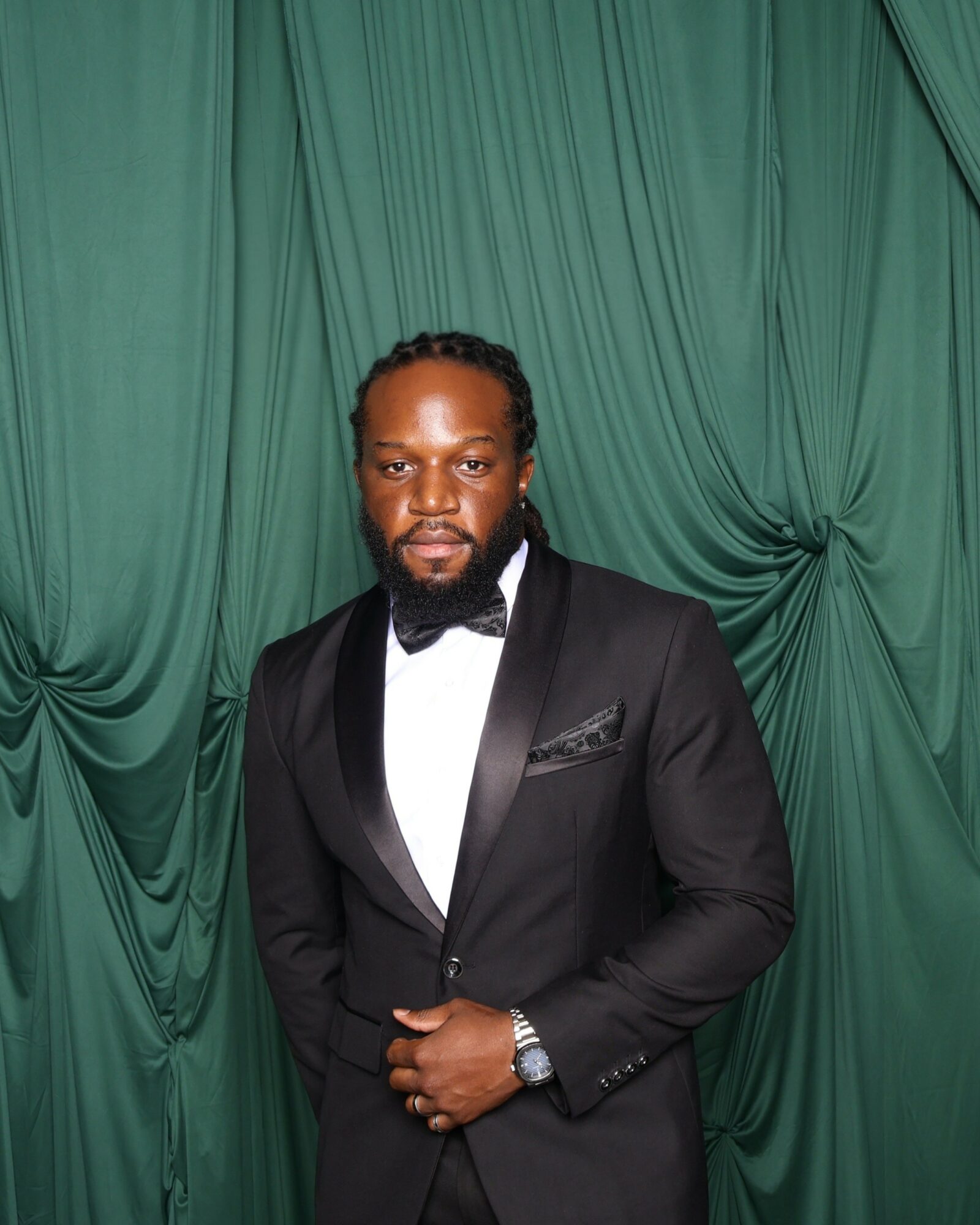 Man in tuxedo with bow tie standing in front of green curtain, looking at camera.