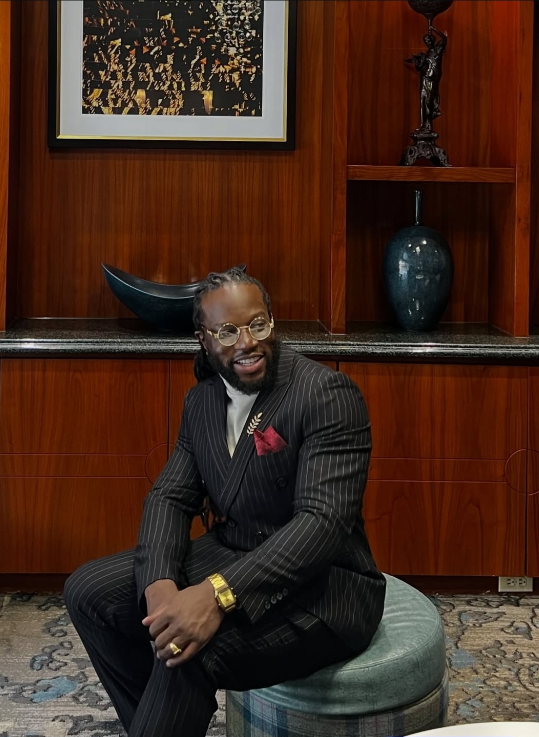 Man in a pinstripe suit sitting on a round ottoman, smiling, in a room with wooden shelves and artwork.