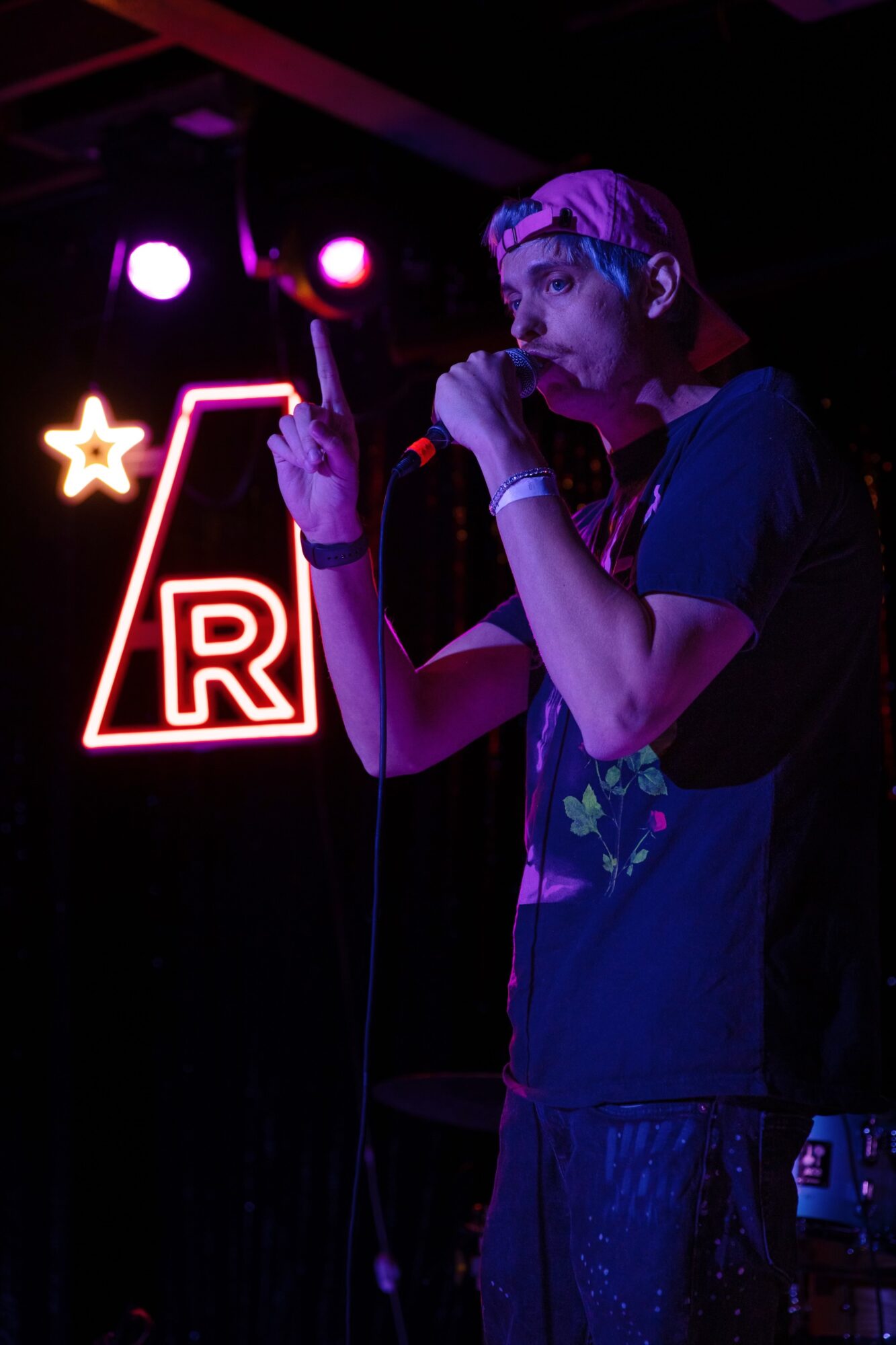 Person with a bandana singing into a microphone on stage, illuminated by purple and pink lights, with a neon sign in the background.