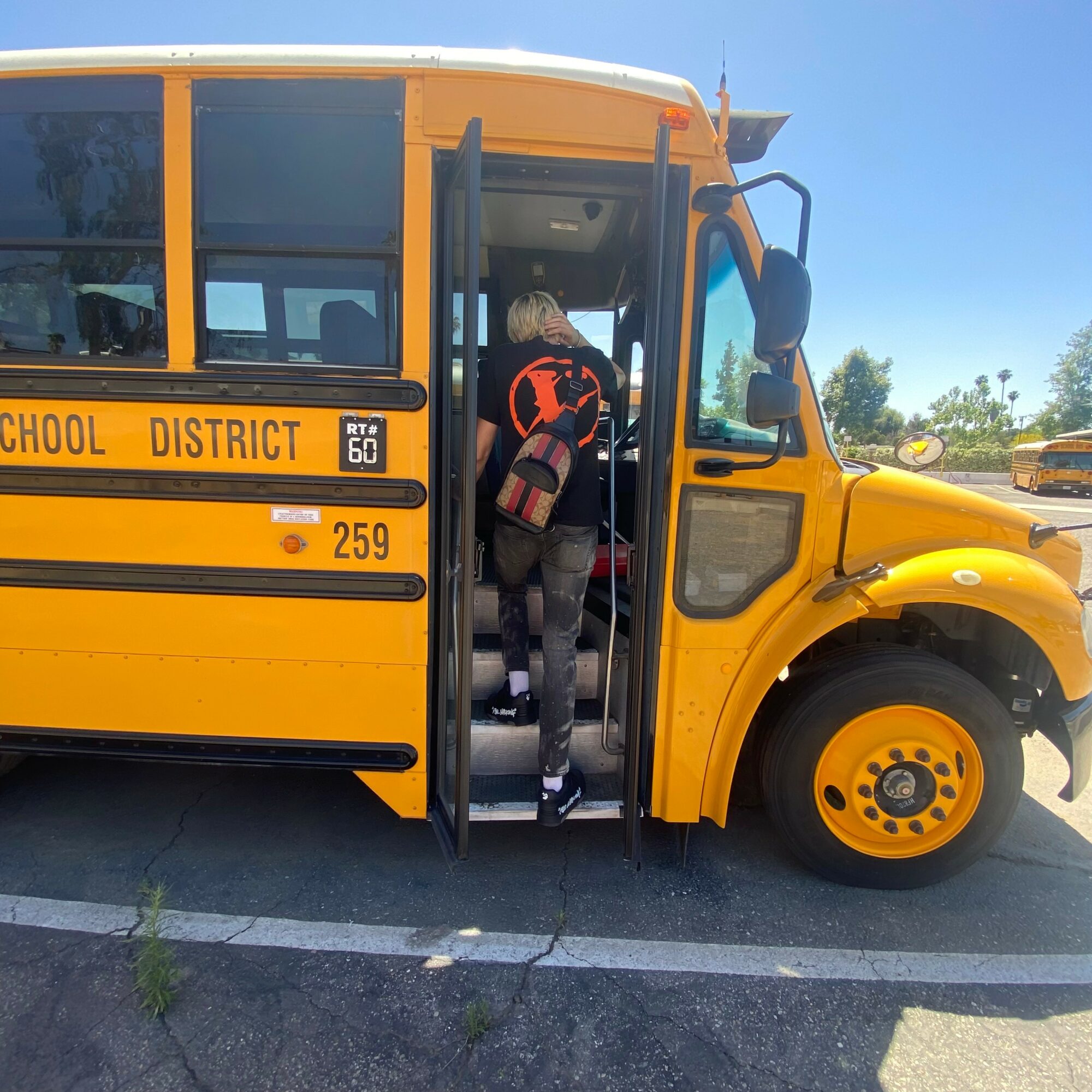 Person boarding a yellow school bus with open door, wearing a backpack, in a parking lot on a sunny day.