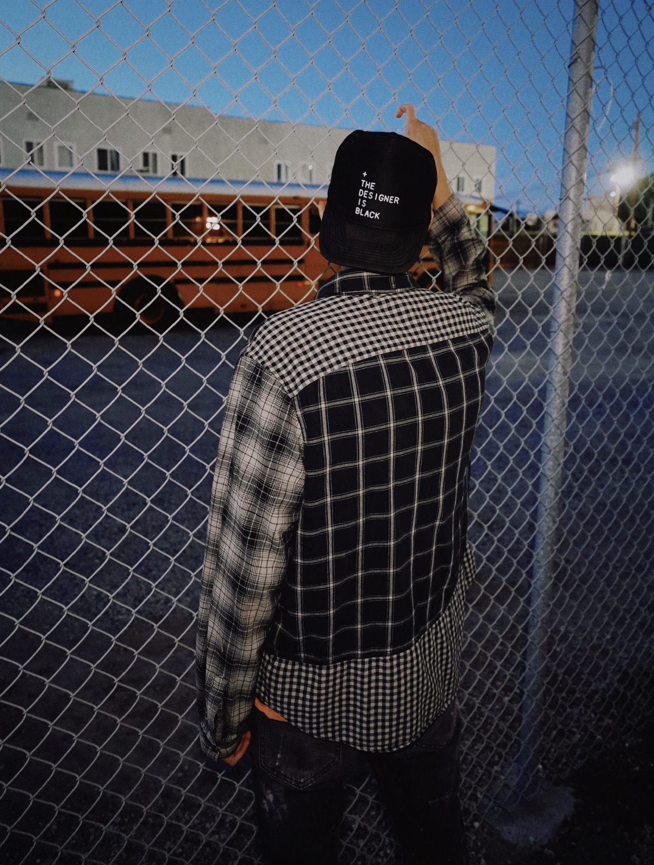 Person wearing a black cap and checkered jacket standing behind a chain-link fence outdoors at dusk.