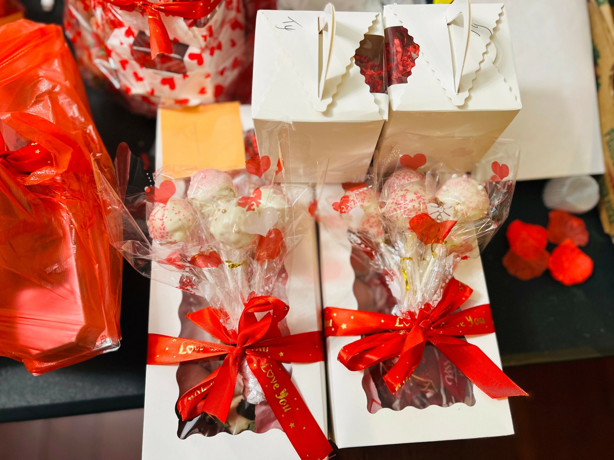 Two gift boxes with flowers and red ribbons, surrounded by other gift items and decorations, on a black surface.