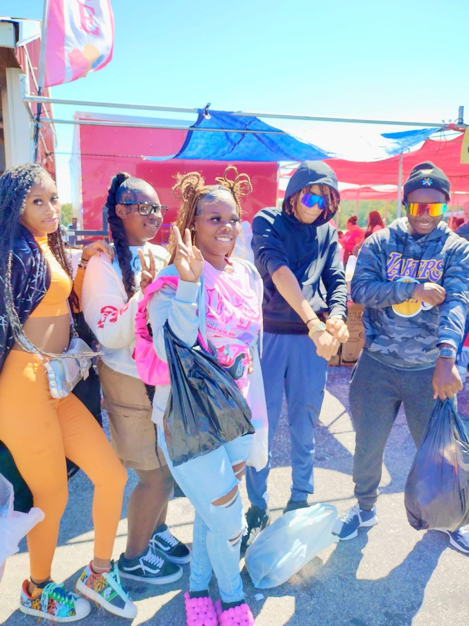 Group of five people standing outdoors, some wearing sunglasses and casual clothing, with tents in the background.