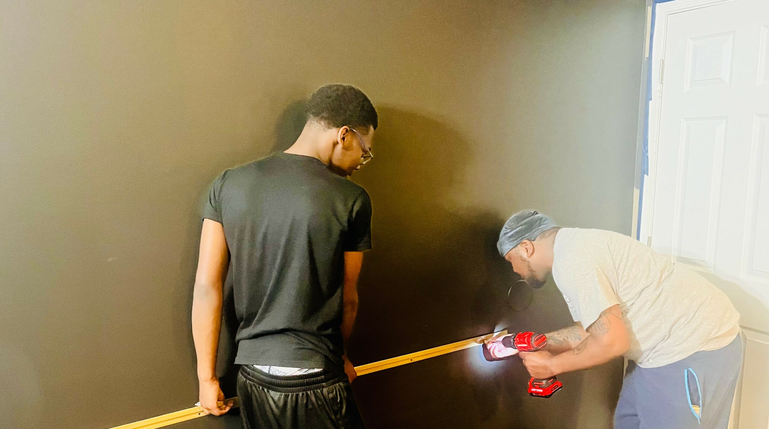 Two people painting a wall, one holding a paint roller and the other using a laser level, in a room with a door.