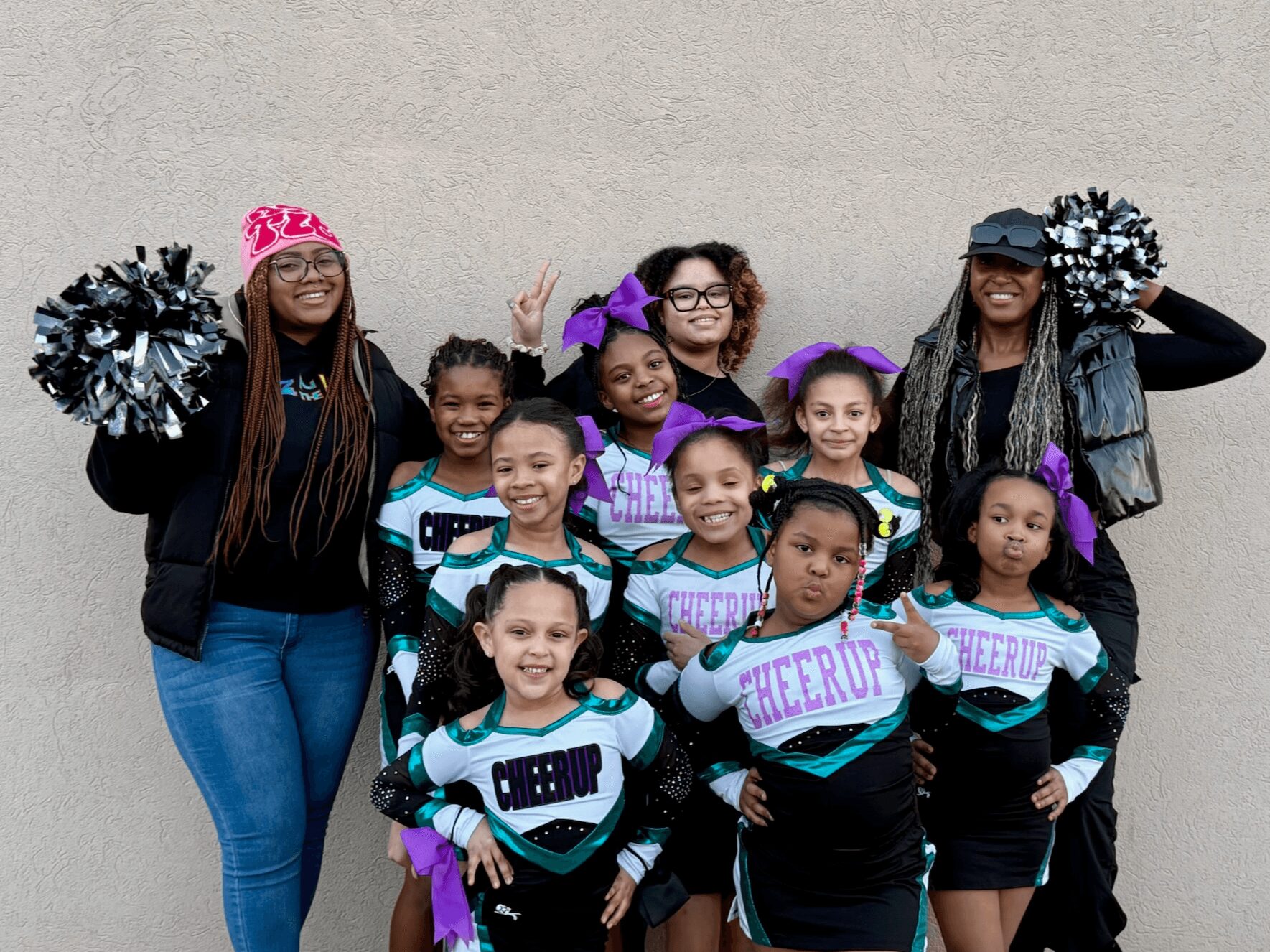 Group of children and adults smiling, wearing cheerleading uniforms and accessories, standing against a plain wall.
