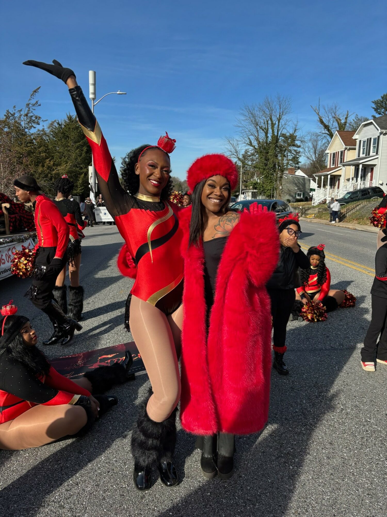 Two women in festive costumes pose outdoors, one in a red and black outfit with arm raised, the other in a red coat and hat, surrounded by people and holiday decorations.