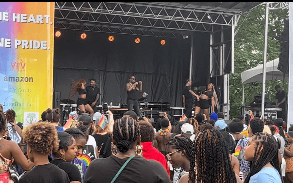 Performers on stage with a crowd watching at an outdoor event, stage with black backdrop, trees in background.