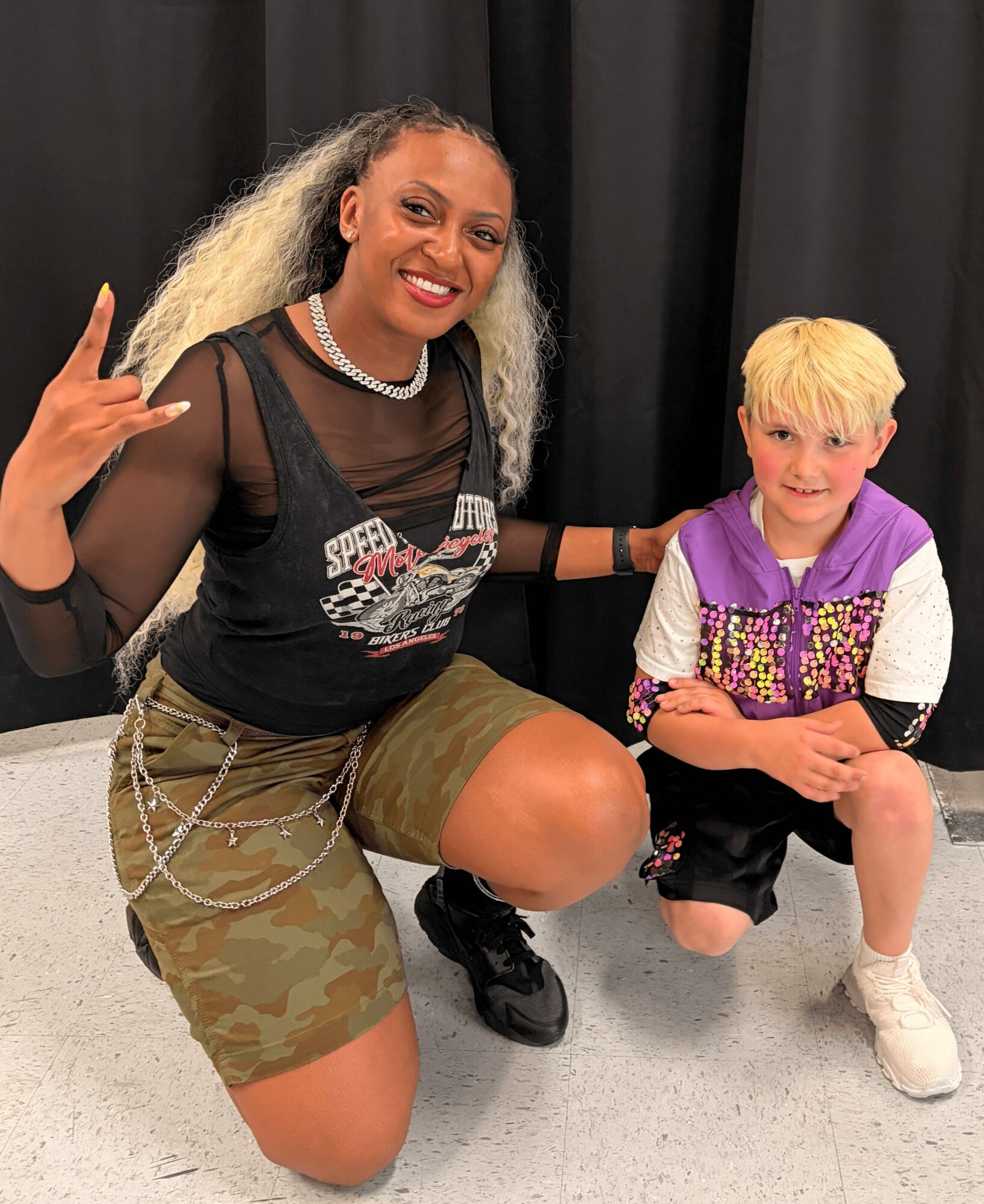 A woman crouches next to a young boy, both smiling, with a black curtain background. Woman makes a rock sign with her hand.