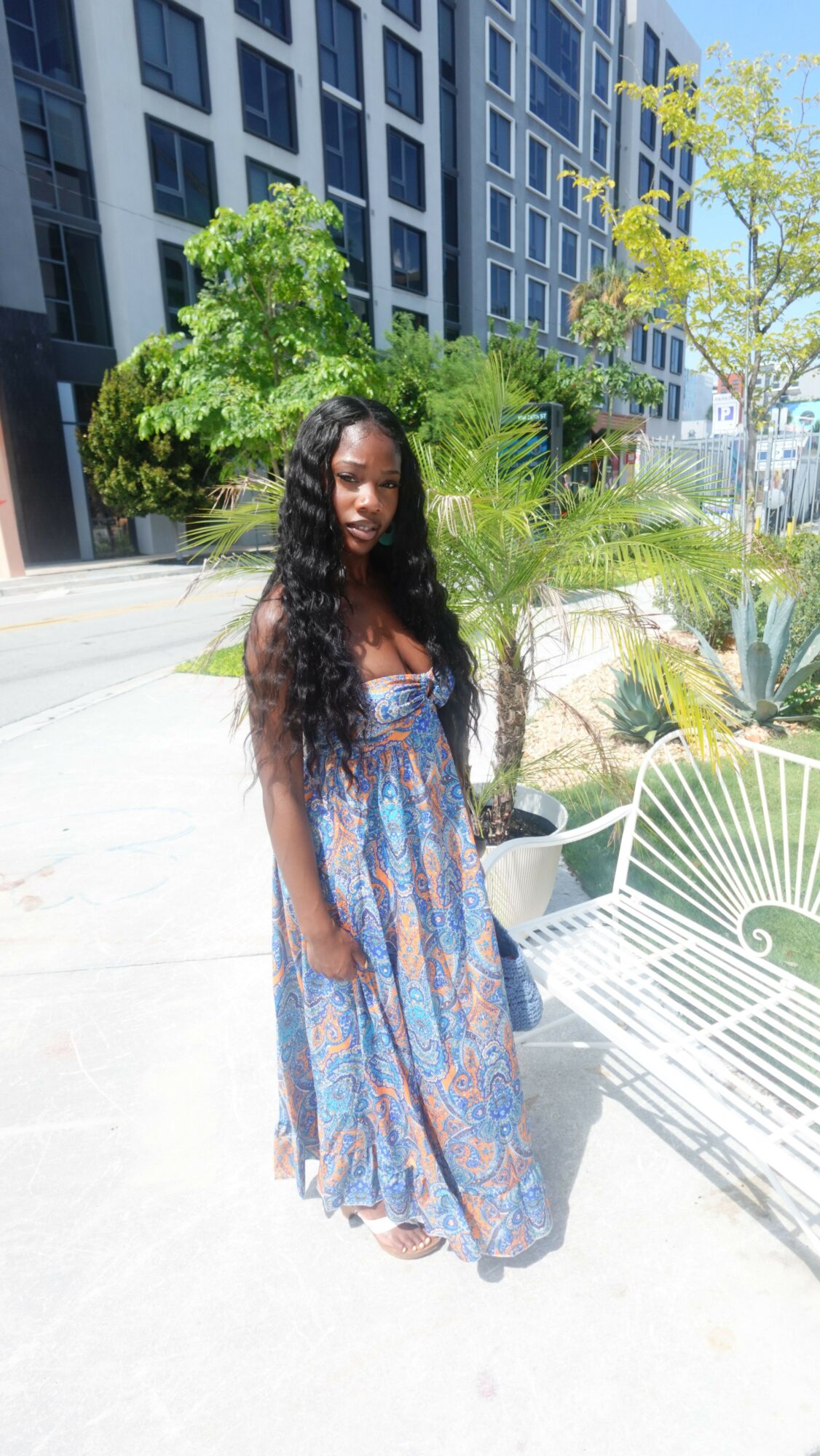 Woman with long hair in a patterned dress standing outdoors near a green tree and modern building.