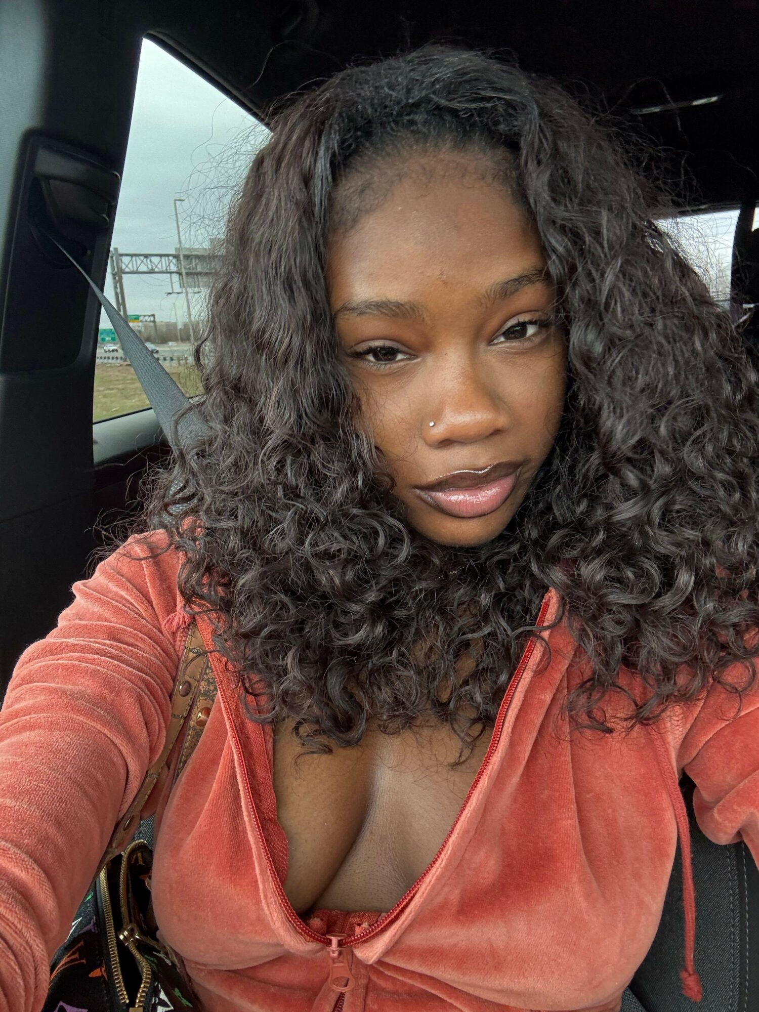 Woman with curly hair wearing a reddish-orange top, sitting in a vehicle, looking at the camera.