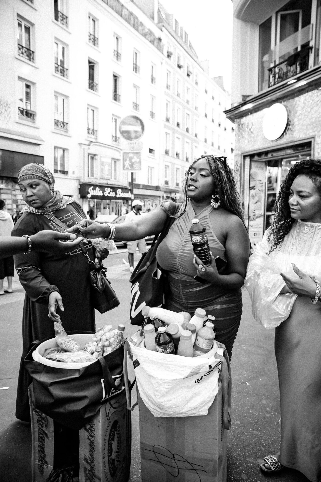 Three women on a city street, one selling drinks from a cart, others browsing and talking, buildings in background.