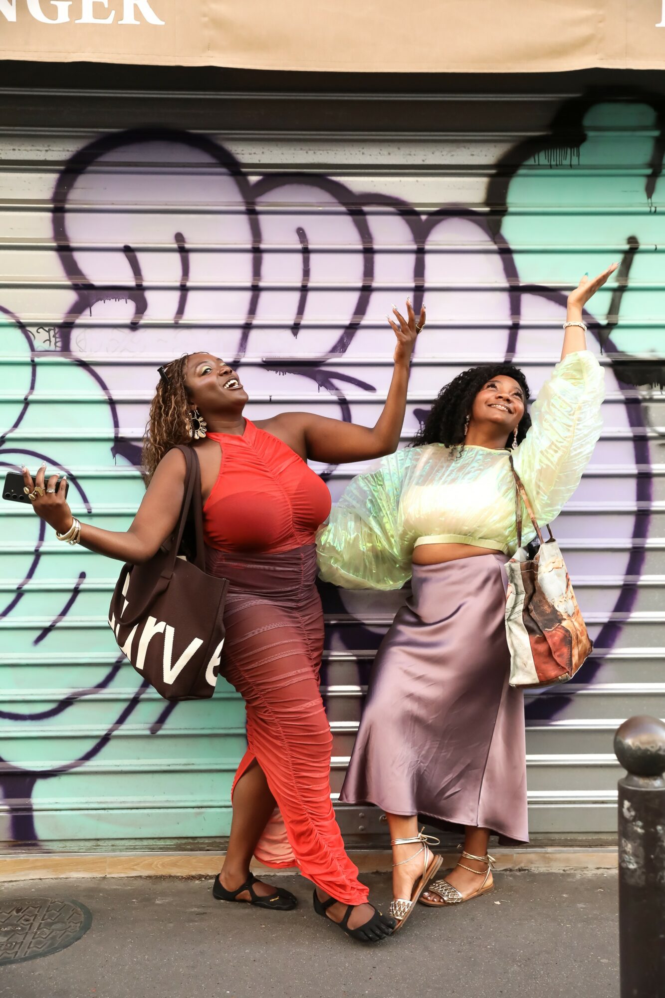 Two women pose in front of graffiti-covered shutter, one with arms raised, wearing colorful outfits, smiling and looking upward.