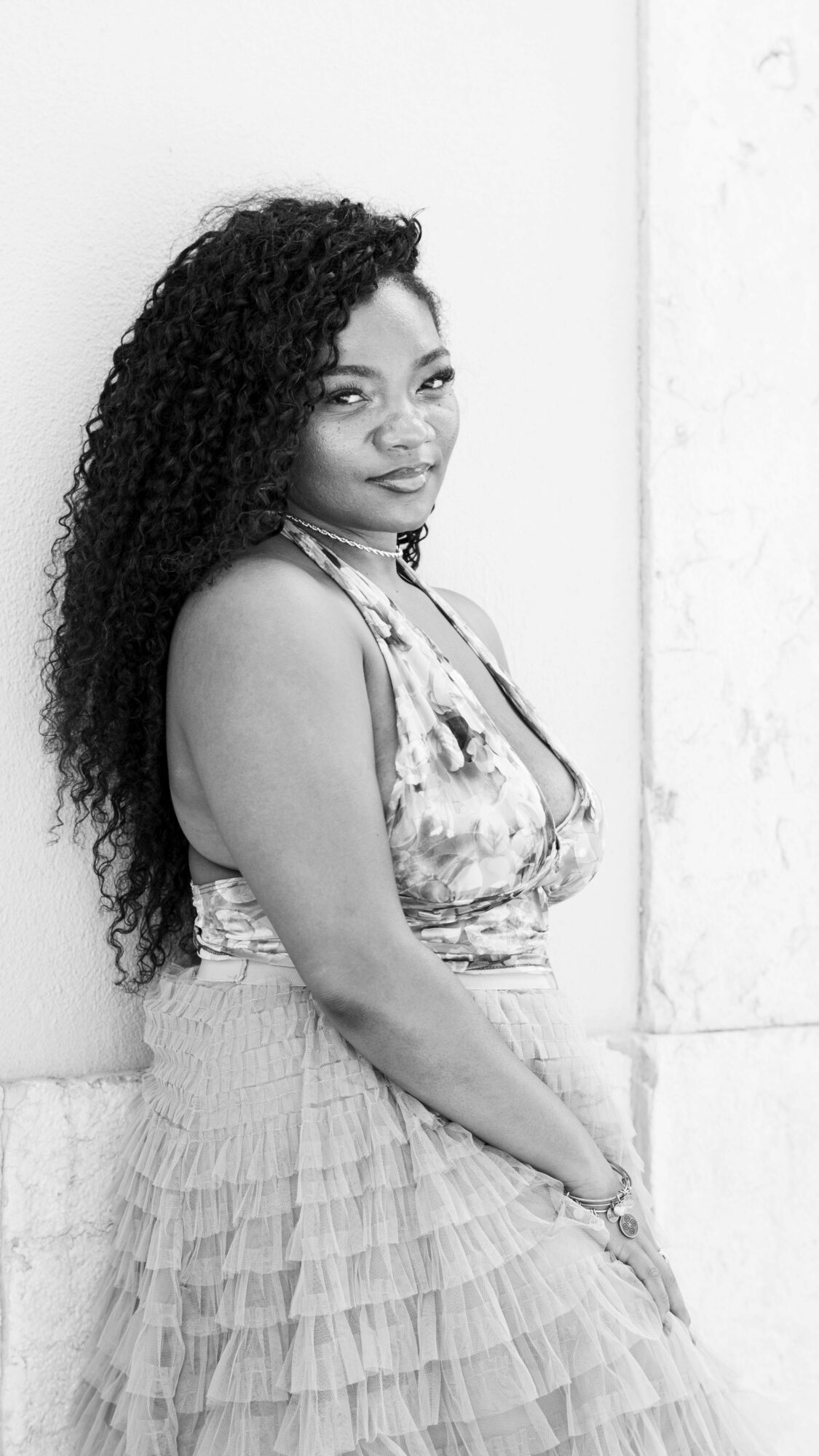 Woman with long curly hair standing against a wall, wearing a patterned top and a layered skirt, smiling.
