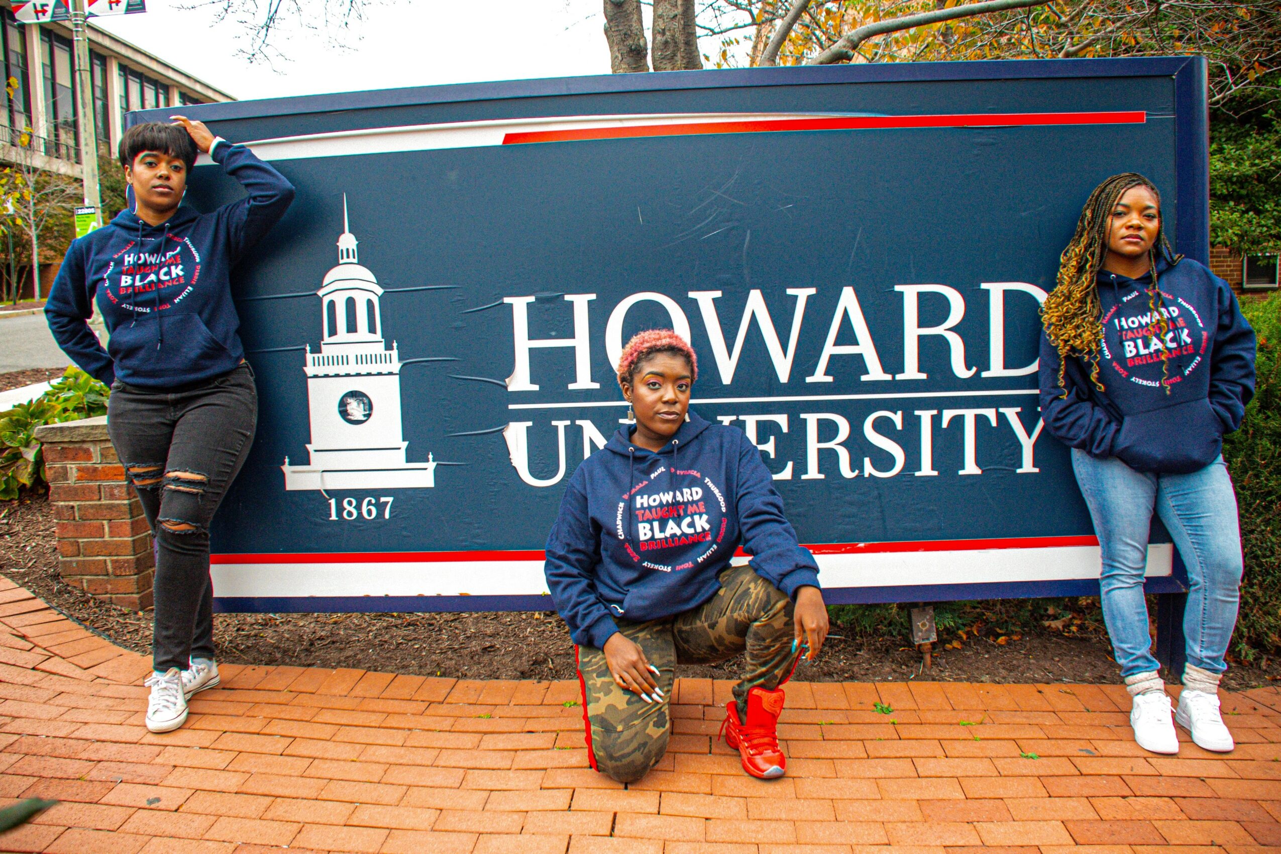 Three students in blue hoodies with Howard University logo pose in front of a Howard University sign, two standing and one kneeling.