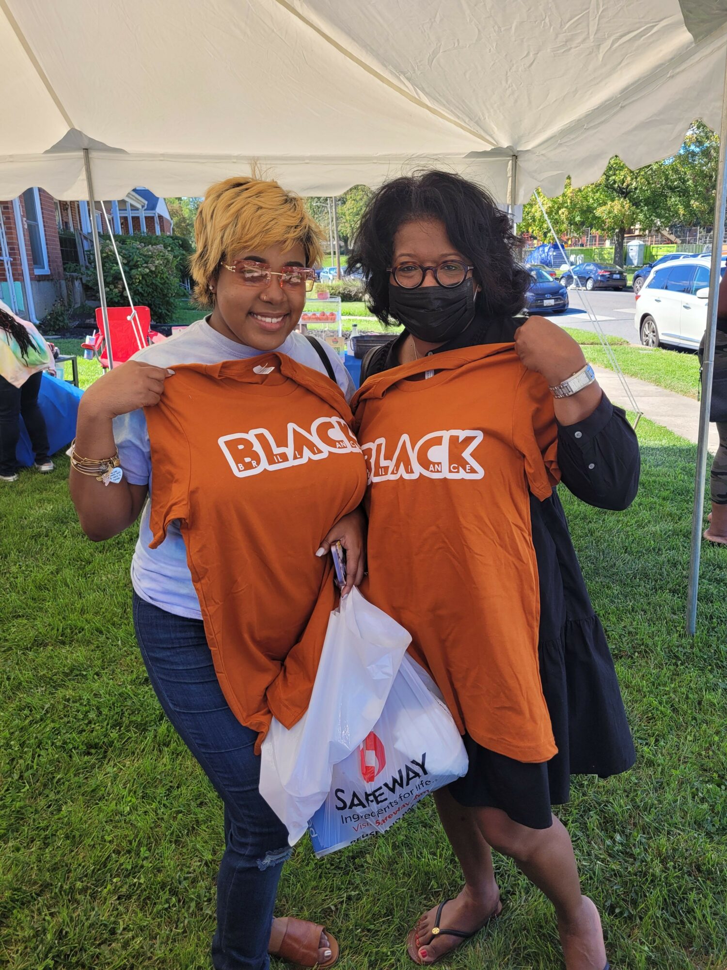 Two women standing outdoors on grass, one with black hair and glasses, the other with blonde hair, both wearing orange shirts.