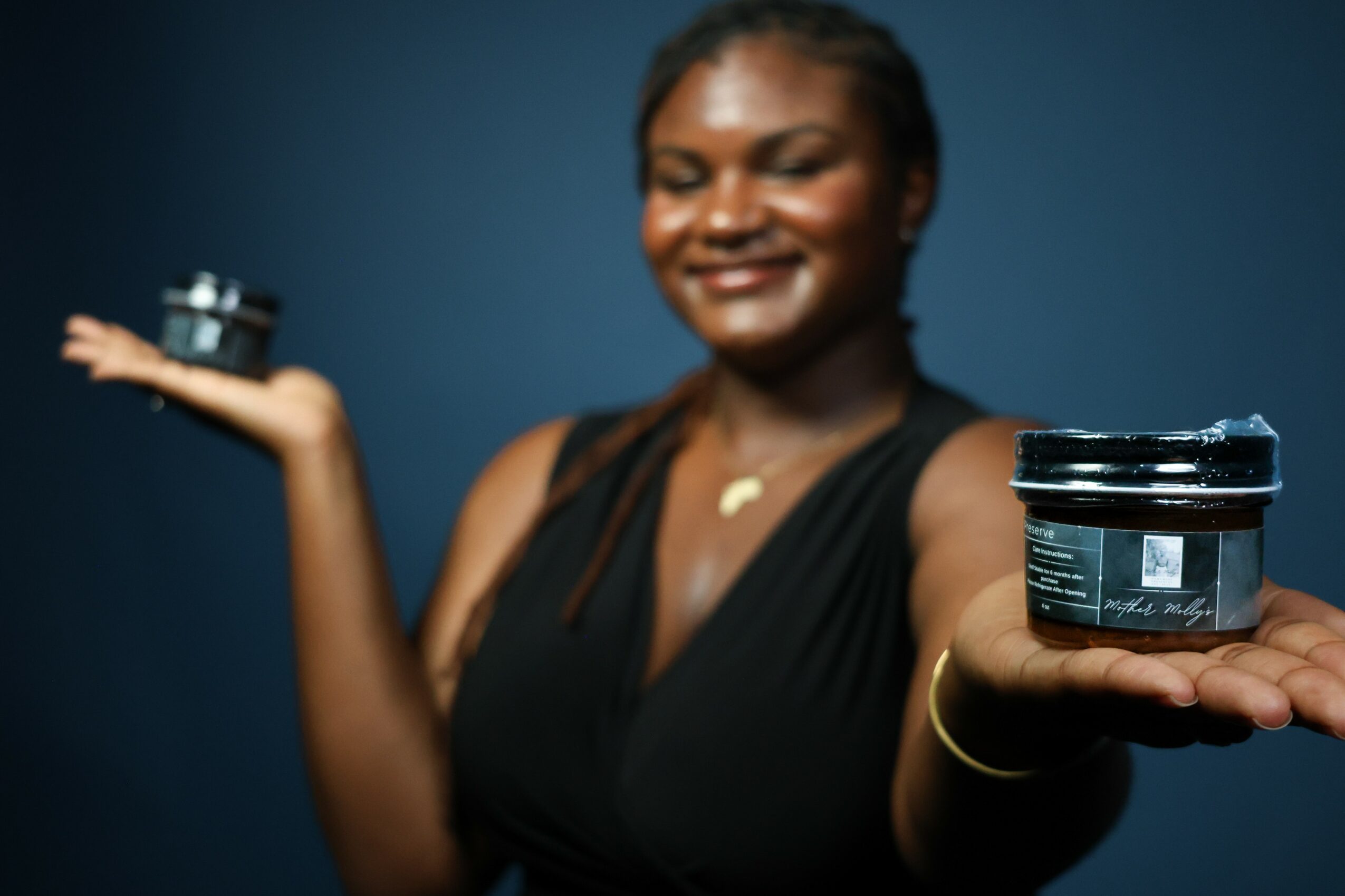Woman smiling holding a small container in each hand, one extended forward and one raised to the side, against a blue background.