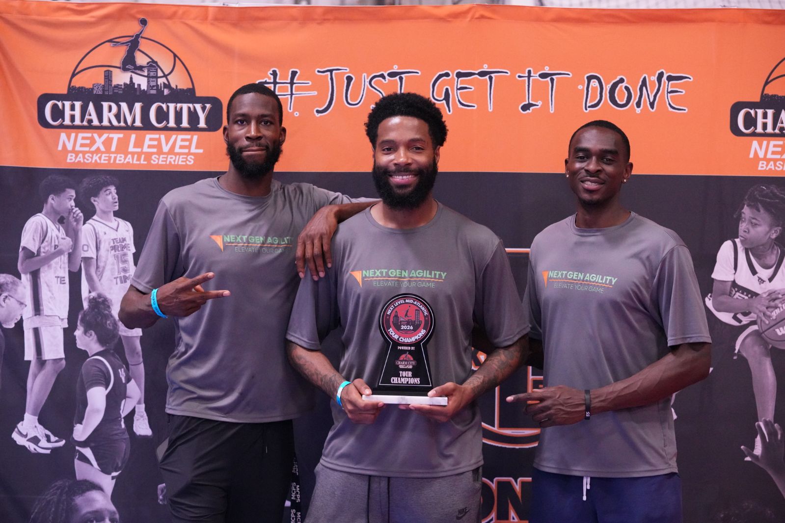 Three men in gray shirts holding a trophy, standing in front of a Charm City basketball banner, smiling.