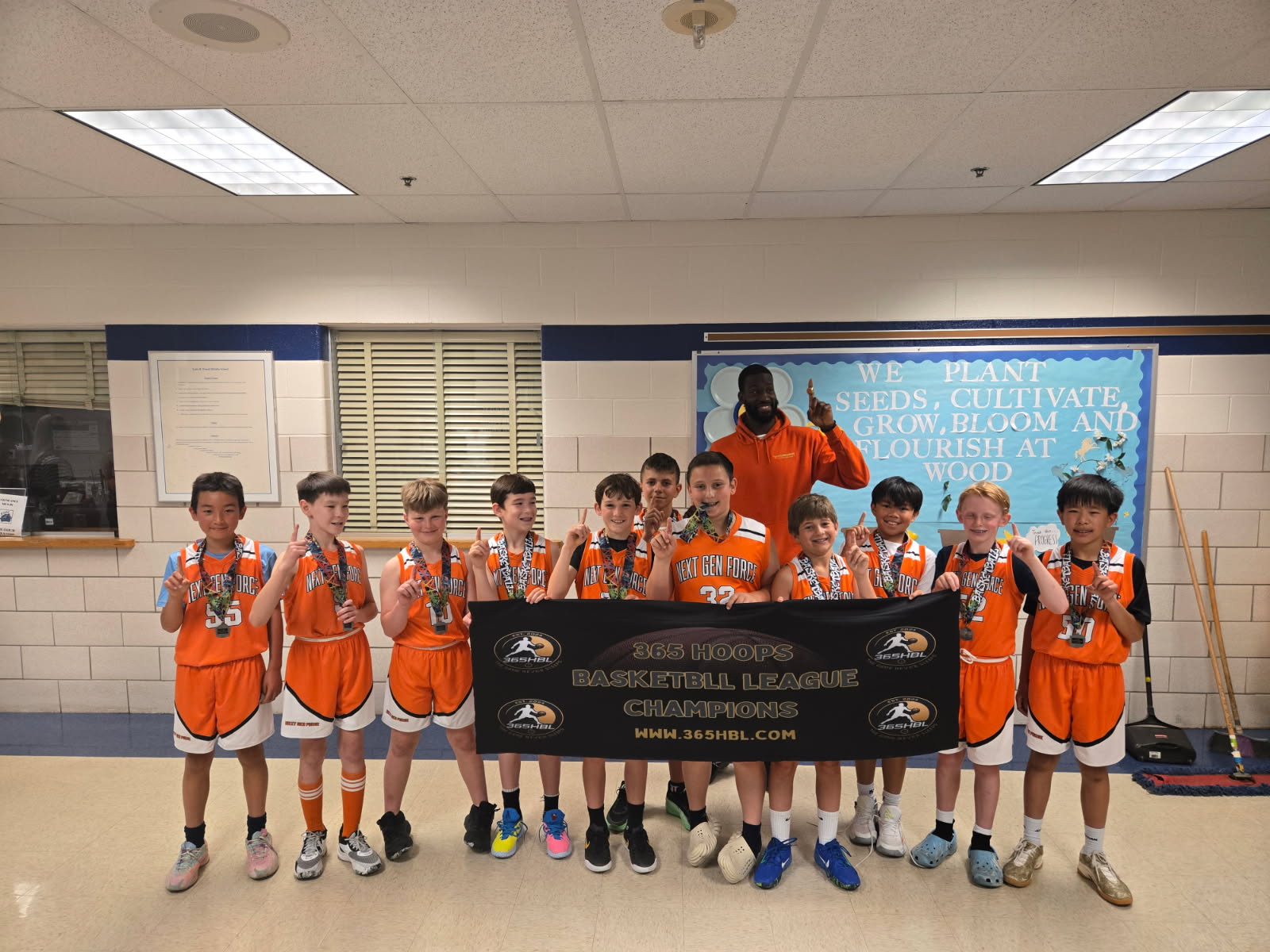 Group of young boys in orange sports uniforms holding a banner indoors, with a blue wall and whiteboard behind them.