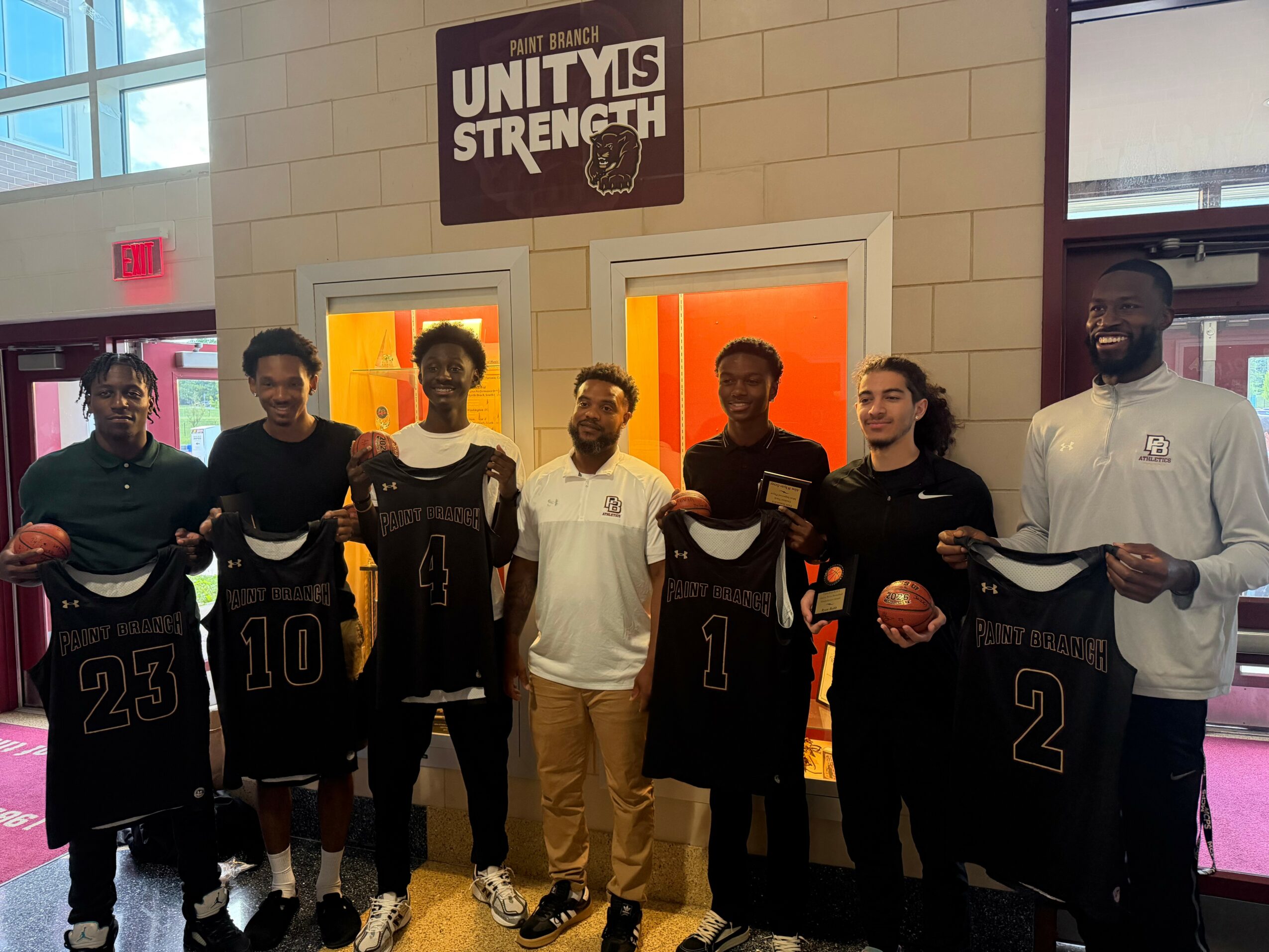 Group of seven people standing indoors, holding sports jerseys and a basketball, with a sign that reads 'Unity is Strength' above them.