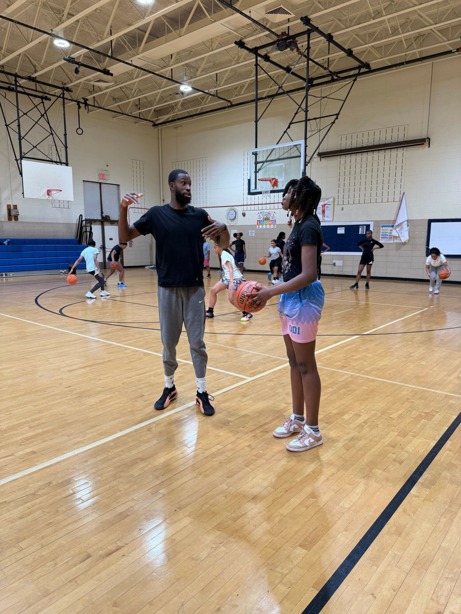 Two women in a gymnasium, one holding a basketball, with others in the background, basketball hoops, and gym equipment.