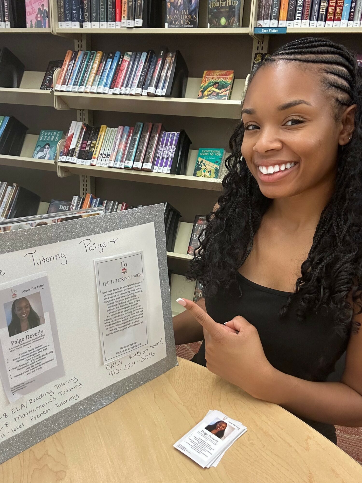 Smiling woman with braided hair points at a framed document on a table in a library, with bookshelves in background.