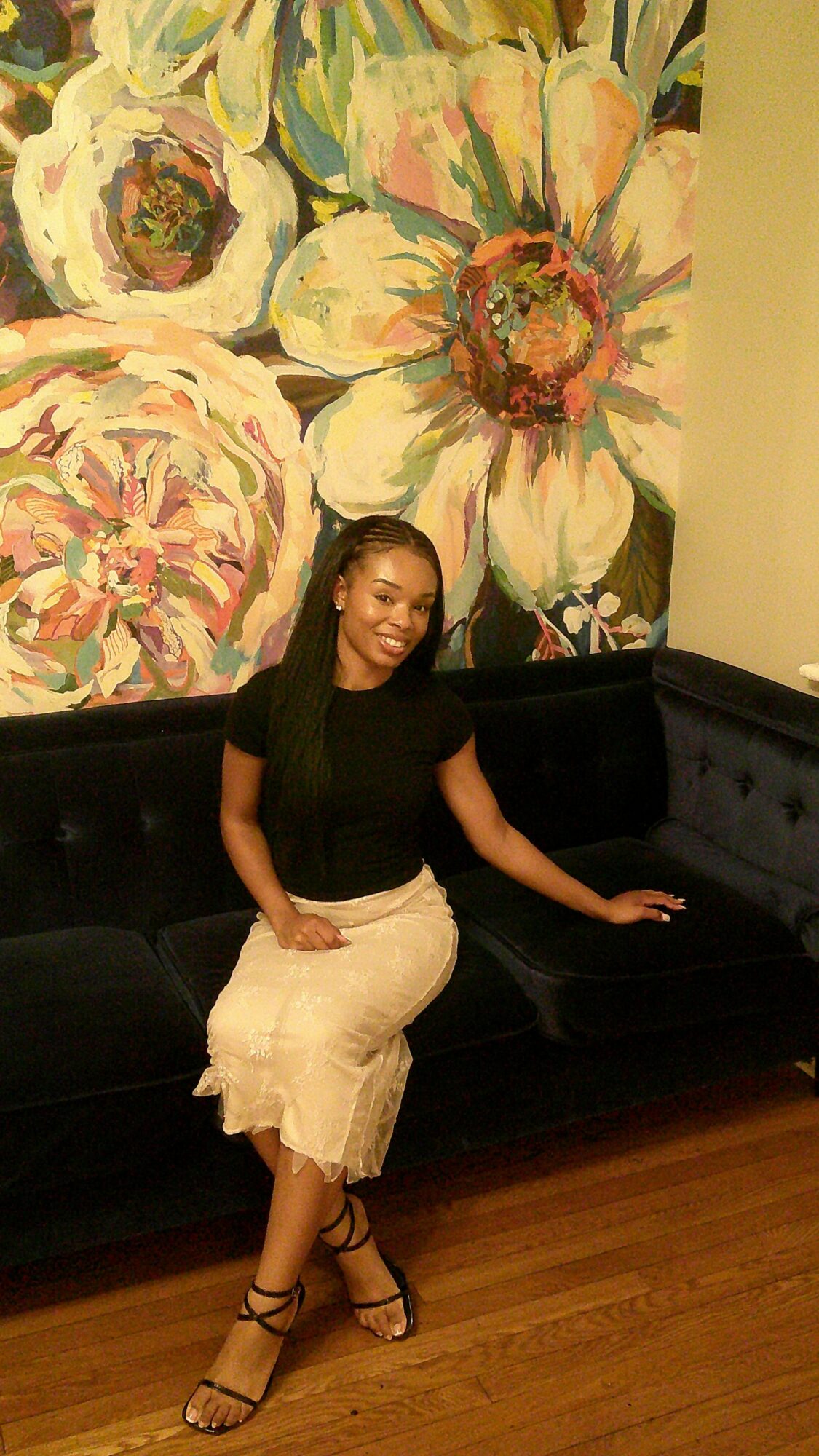 Woman standing next to a floral mural, wearing a black top and light-colored skirt, with one arm raised and smiling.