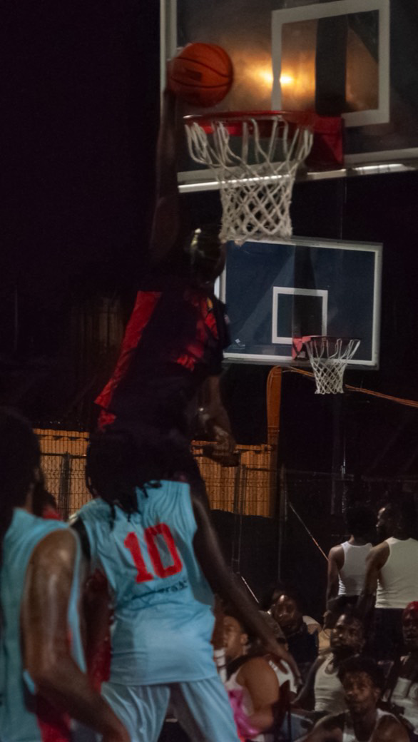 Person in red and black shirt jumps near basketball hoop at night, with other players and spectators nearby.