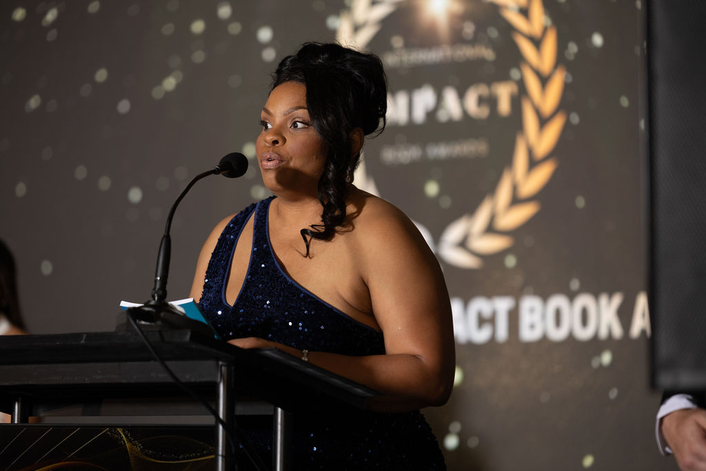 Woman in a dark blue dress speaking at a podium during an event, with a backdrop featuring a laurel wreath and text.